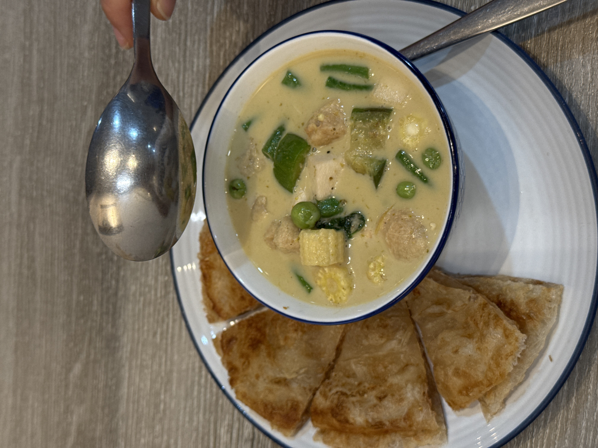 A close-up overhead shot shows a bowl of green curry with chicken, peas, and corn on a white plate with a blue rim. The curry is a creamy light green color, and the chicken is cut into bite-sized pieces. Several pieces of crispy fried flatbread, possibly roti, are arranged around the bowl. A metal spoon is being held over the bowl, as if about to scoop some curry. The plate is on a wooden table, which appears to be a common surface found in many restaurants in Bangkok, Thailand, such as those located in the Sukhumvit area, known for its diverse culinary scene. The lighting suggests an indoor setting, possibly a restaurant or a home kitchen.