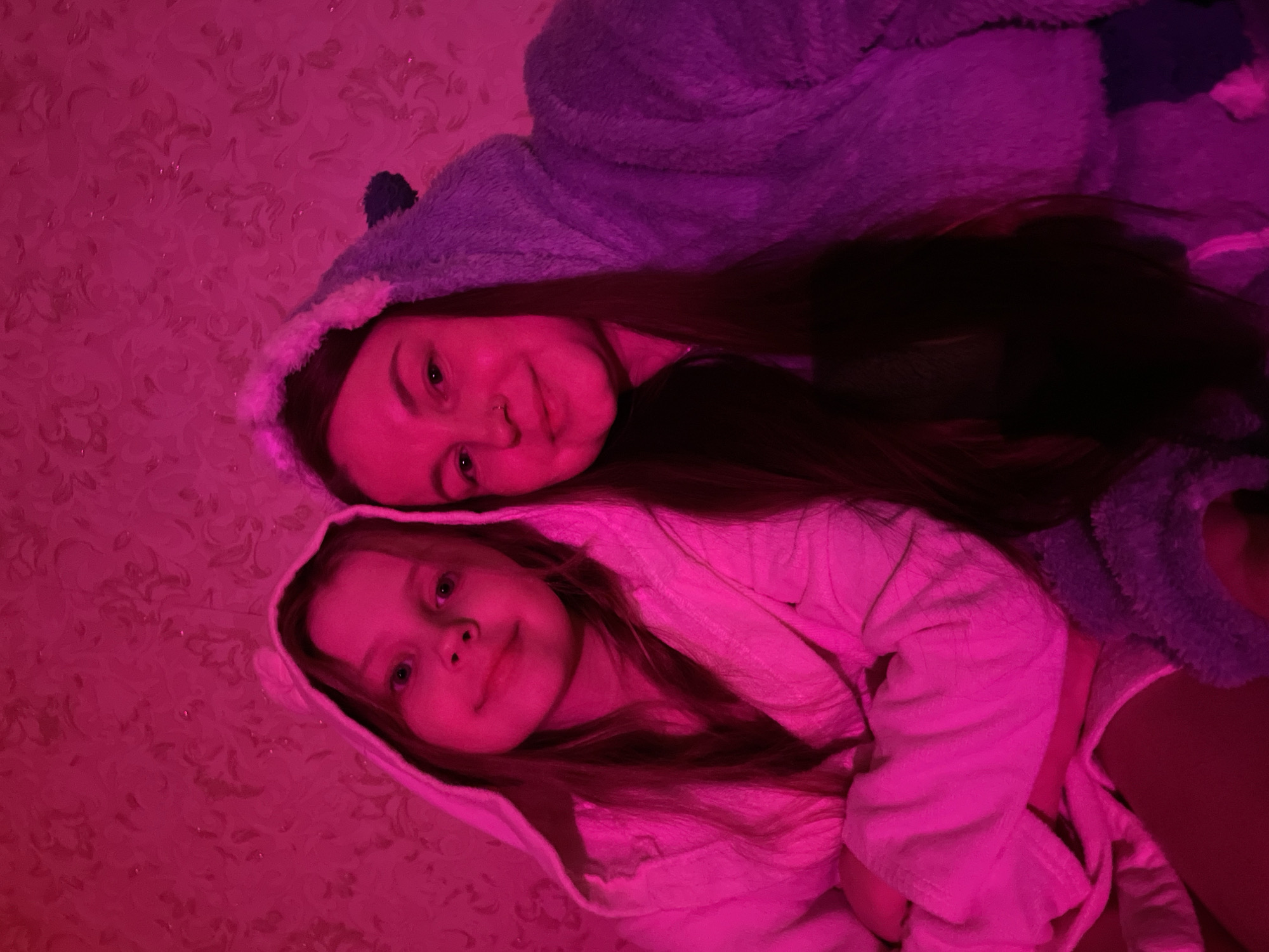 The image captures two females, an adult and a young girl, lying together in an indoor setting, likely a bedroom, in Izhevsk, Russia. Both subjects are looking towards the camera with relaxed and gentle expressions. The adult wears a purple hooded robe, possibly an animal-themed onesie with a dark ear visible, and has long dark hair and a small nose piercing. The young girl is dressed in a pink hooded robe and has lighter, long hair.
The entire scene is illuminated by a strong, artificial pink or magenta light source, giving the image a distinctive, vibrant hue. This lighting casts a deep pink glow on their skin, clothing, and the background, which features a subtly patterned wallpaper. The intense artificial light suggests the photograph was taken in a dimly lit room or at night, creating a cozy and intimate atmosphere. No text is visible in the image.