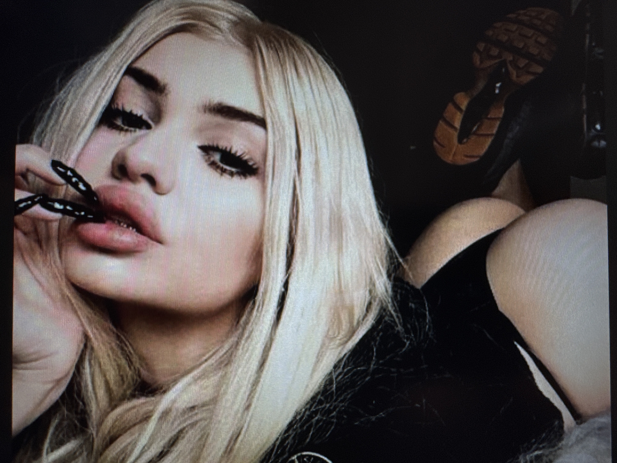 A dimly lit portrait shows a person, likely female, with long, straight blonde hair framing her face. She has pronounced dark eyebrows, heavy black eyeliner, and long, dark eyelashes. Her full lips are slightly parted in a pouting expression, and her left hand, adorned with long, black, textured nails, is held close to her mouth. She is wearing a dark, likely black, garment, with a subtle metallic detail visible near the bottom center of the frame.
In the upper right background, a distinctive shoe is visible, featuring a thick, patterned sole in shades of brown and black, suggesting a platform or high-heeled boot. The background is predominantly dark, indicating an indoor setting with artificial or low lighting, drawing focus to the subject. A curved, skin-toned area in the lower right suggests the outline of a hip or thigh, indicating a reclining pose. The image has a slightly grainy or pixelated quality. The overall mood is confident and alluring. No text is visible. This image is from Varna, Bulgaria.