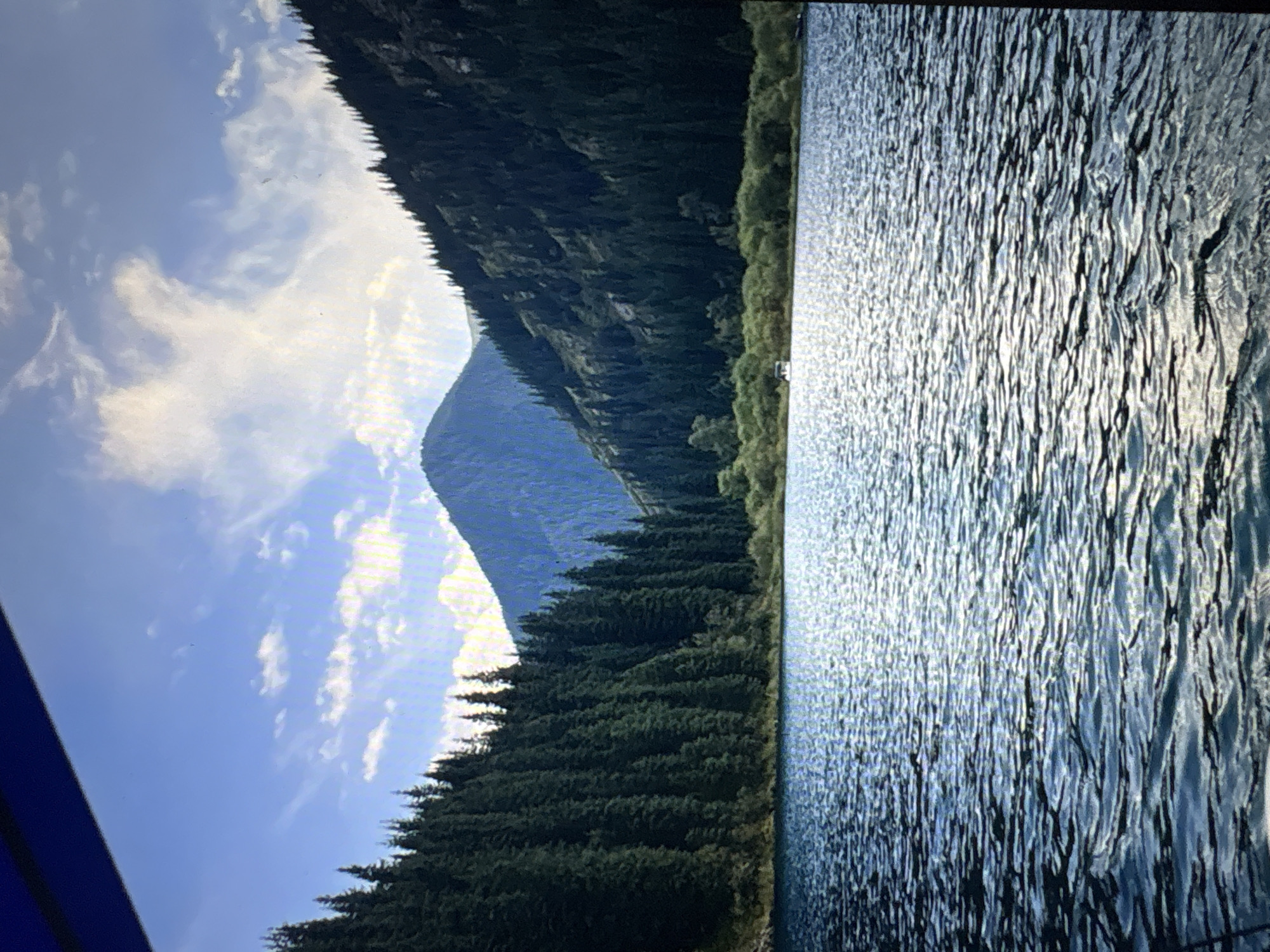 A landscape scene shows a body of water in the foreground, reflecting light that creates vertical striations in the water. The right edge of the water is bordered by a line of green trees. Further to the left, a forested mountain slope rises sharply. A taller, blue-tinged peak is visible behind this slope. The sky above features a mix of light blue and white, with scattered cumulus clouds. The image is taken outdoors, with sunlight affecting the water and illuminating the sky. The image is slightly rotated.
Mława, Poland is not visible in this image.