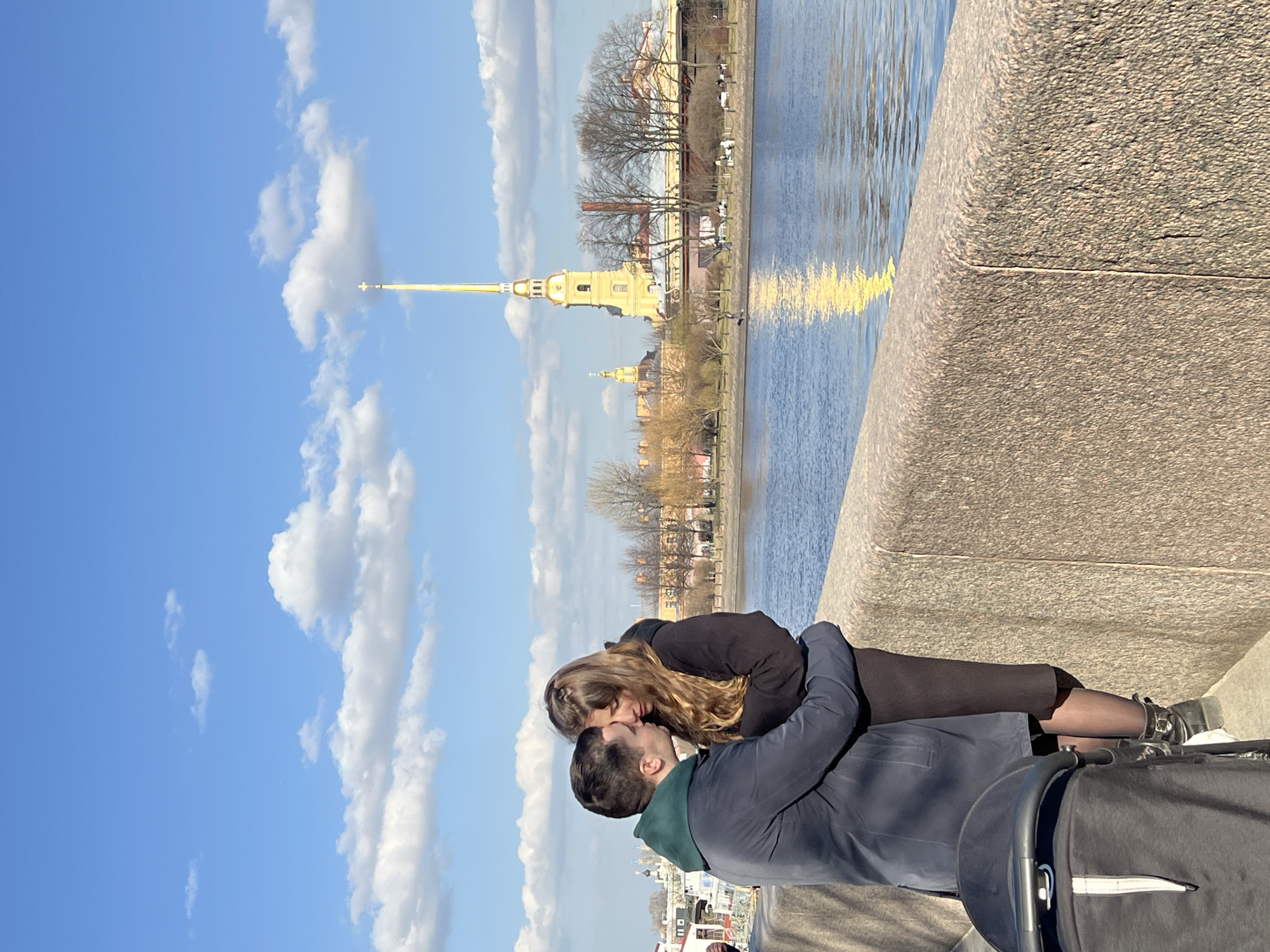 The image displays an outdoor daytime scene in Saint Petersburg, Russia, under a clear blue sky with scattered white cumulus clouds. In the immediate foreground, a textured, light brown granite embankment wall spans the lower portion of the frame. To the left, the top portion of a dark-colored baby stroller with a visible white accent strip is present.
Two individuals, a man and a woman, stand closely together against the embankment wall, engaged in an intimate interaction. The man, positioned on the left, wears a dark blue or grey coat over a green hooded sweatshirt. His right arm is around the woman's back, and his head is bowed towards hers, with their foreheads touching or his head resting on her temple. The woman, on the right, has long, wavy brown hair and wears a dark coat or dress, dark hosiery, and dark boots with visible buckles. Her eyes appear closed or downcast as she leans into the man.
Beyond the embankment, the Neva River flows, its blue surface reflecting a prominent vertical streak of golden light. On the opposite bank of the river, the Peter and Paul Fortress complex is visible. Its yellow-ochre buildings, including the iconic Peter and Paul Cathedral, are prominent. The cathedral's tall, slender golden spire, topped with a cross, rises distinctly into the sky, reflecting the bright sunlight. Bare trees line the distant bank, indicating either late winter or early spring conditions. The scene captures a moment of personal connection against the backdrop of a significant city landmark.