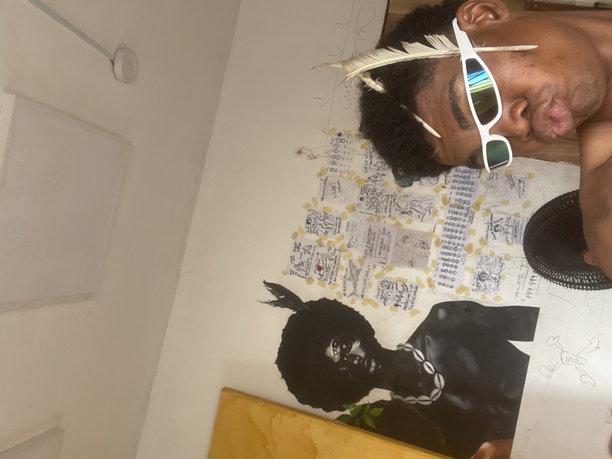 The image captures a selfie taken indoors, likely in a room in Ado, Nigeria, given the context. The subject, a person with dark, curly hair adorned with two light-colored feathers and a small, twig-like object, is the focal point. They are wearing sleek, white, futuristic sunglasses with mirrored lenses that reflect a rainbow of colors. Their lips are pursed, creating a playful or provocative expression.
Behind the subject, the wall is covered with a collage of papers, taped haphazardly. These papers feature a mix of drawings, handwritten text, and what appear to be diagrams or flowcharts. To the left of the subject is a large, black-and-white painting of a person with an afro, wearing a shell necklace. The painting is set against a bright yellow background. The overall impression is one of a creative or artistic space, with personal touches and a sense of organized chaos. The lighting appears to be artificial and originates from the ceiling, indicated by the presence of two square light fixtures. A black fan is partially visible in the foreground on the right side of the image. The wall also has some rudimentary drawings, including a skull and crossbones.