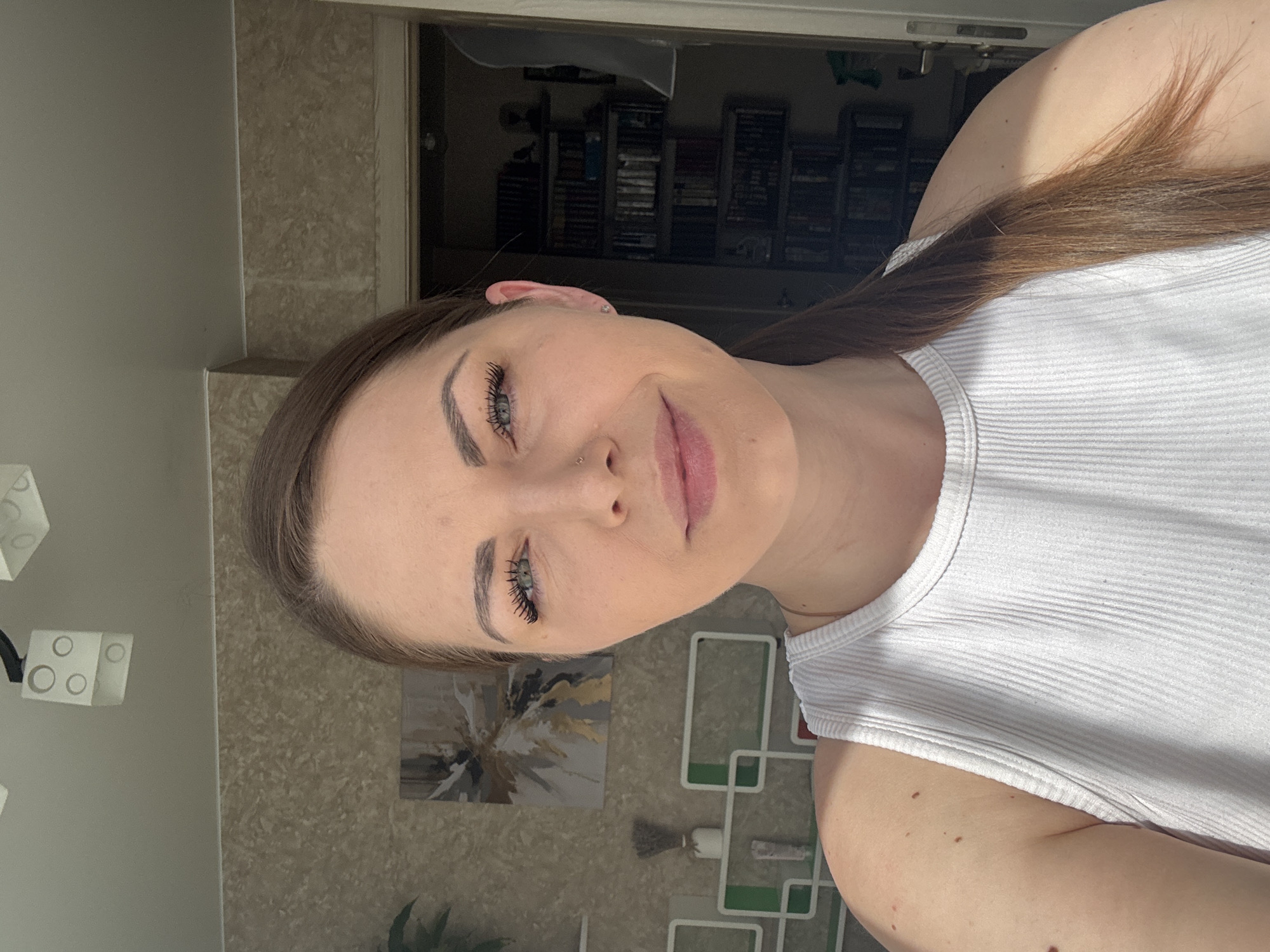 A woman with fair skin, light brown hair pulled back into a ponytail, and wearing a white ribbed tank top, is seen in a close-up, slightly rotated portrait. She has well-defined eyebrows, noticeable eyelashes, and a piercing in her nostril.
Behind her, a doorway leads into a dimly lit room with a wall lined with what appear to be numerous VHS tapes. In the background, on the wall, there is an abstract artwork with gold and gray hues. A white geometric sculpture, possibly a wall decoration, is visible in the lower right, as well as a few green leaves from a plant. The location is in the city of Unknown, Unknown.