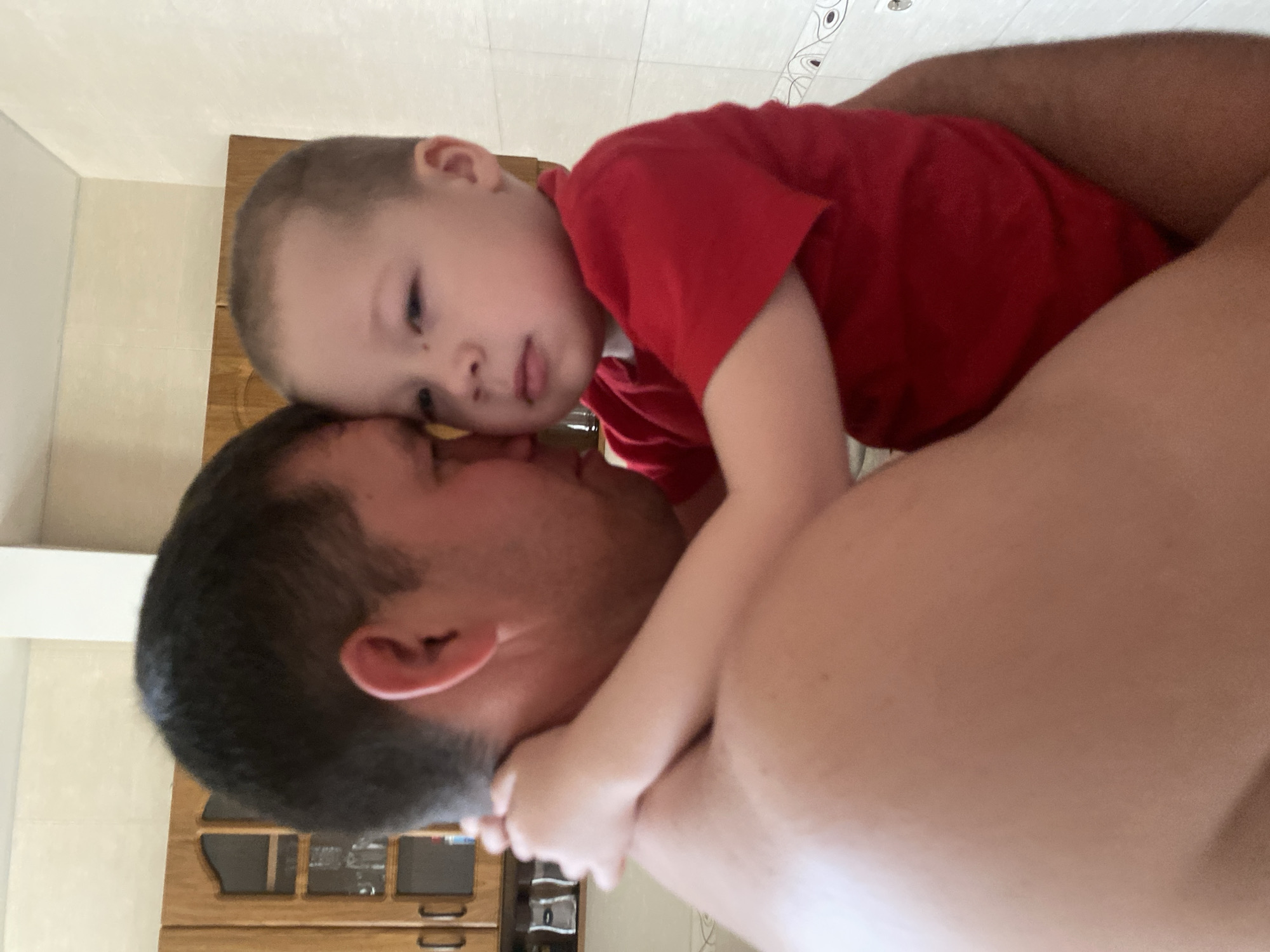 A close-up indoor shot depicts a child in a red shirt leaning against a man. The man has short, dark hair and the child has fair skin. The child's arms are around the man's neck. In the background, there's a wooden cabinet with drawers and a white wall. The location of the scene is not determinable from this image, including if it is in Sochi, Russia.