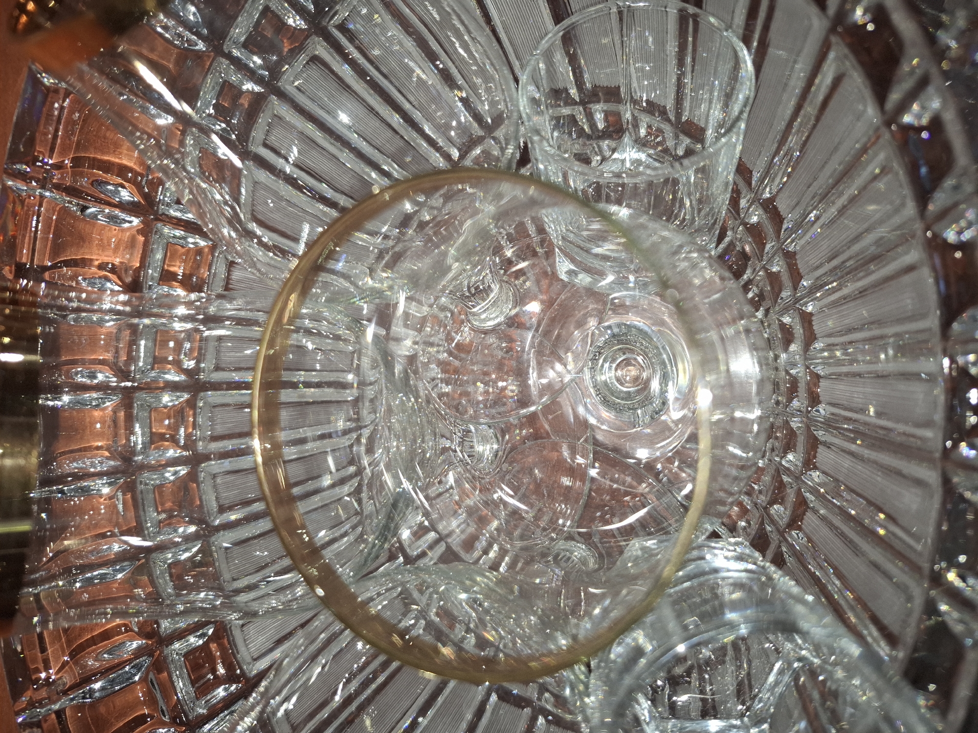 The image shows a close-up, top-down view of a crystal bowl with intricate geometric cuts and a gold rim. Inside the bowl, several clear glass goblets are nested together, some appearing to be champagne flutes. The lighting creates reflections and refractions of light throughout the glass surfaces. The background is a textured, reddish-brown surface that appears to be a table or countertop. There is no identifiable information in the image that would allow for the determination of a specific location in Almaty, Kazakhstan.
