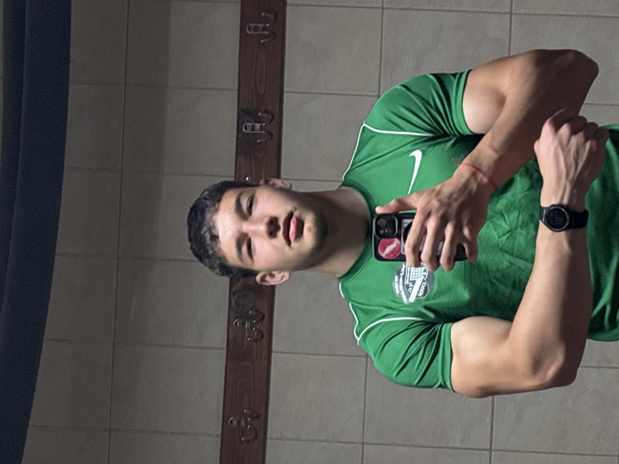 A young man is posing for a selfie in a locker room with tiled walls. He is wearing a green athletic shirt with white trim and a Nike logo, and a black smartwatch on his left wrist. He is holding a cell phone in his right hand. Behind him, there is a dark wooden rack with metal hooks attached to the tiled wall. The scene is likely in Zlatograd, Bulgaria, due to the reference in the prompt.