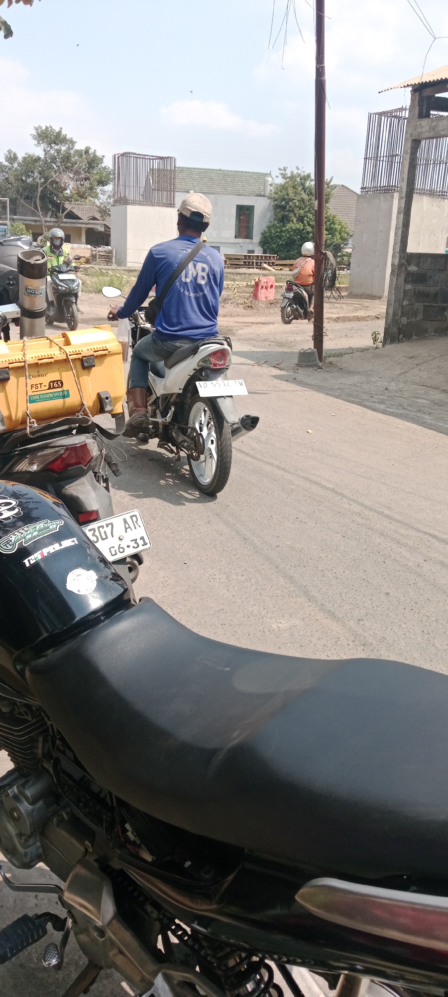 The image captures a scene on a street in Purwomartani, Indonesia, likely during daytime given the bright sunlight and clear sky. In the foreground, the seat and rear section of a black motorcycle are visible. Attached to the rear of another motorcycle, a large yellow box labeled "FST-16S" and "CORE FUSION SPLITER" is prominently displayed. A man wearing a blue long-sleeved shirt and brown boots is seated on a white motorcycle, facing away from the camera, with a black bag slung across his shoulder. The motorcycle has a license plate that reads "AD 5532 TW". To the left, another motorcycle is parked, and a person in a green shirt and helmet is also on a motorcycle. In the background, a person in an orange shirt is seated on a motorcycle. The environment features a paved road, some greenery with trees, and buildings with white walls and green roofs, including what appears to be a construction site with scaffolding and concrete structures. The overall impression is of a street scene with multiple motorcycles and individuals, possibly related to some form of delivery or construction work.