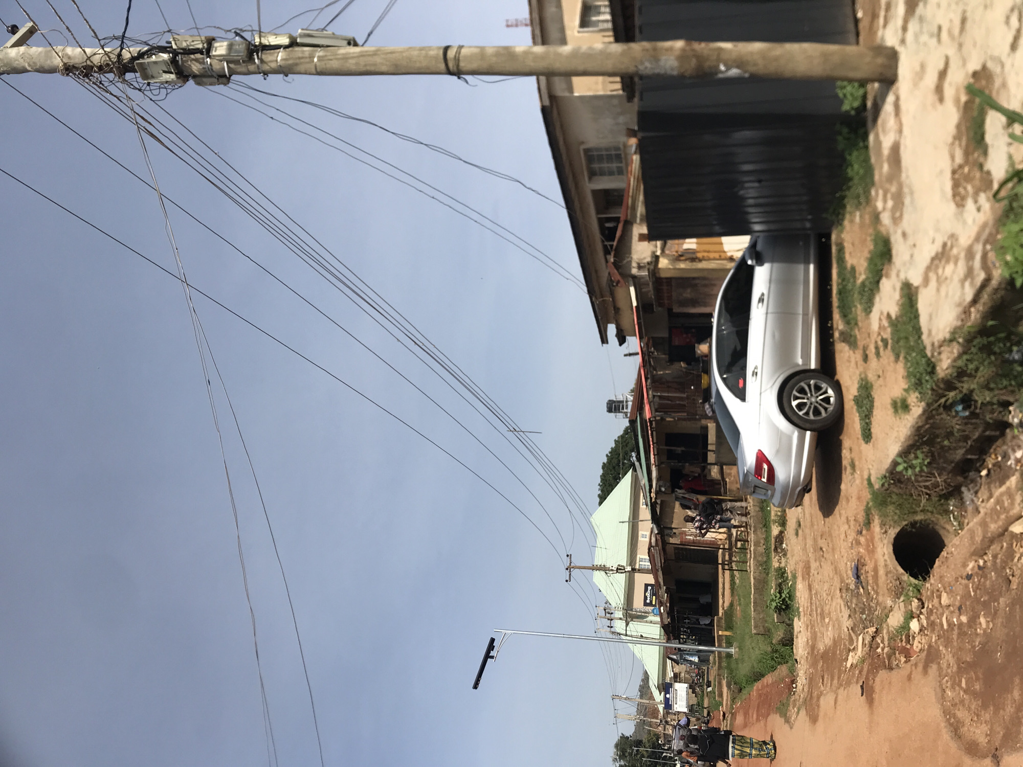 The image captures a street scene in what appears to be an urban or semi-urban area in New Stadium, Nigeria, under a bright, clear daytime sky. The primary subject is a utility pole heavily laden with numerous overhead wires, some of which appear tangled or loosely strung. This pole stands prominently against the sky on the left side of the frame.
To the right, a white sedan, possibly a Mercedes-Benz given its wheel design, is parked alongside a row of buildings. The car's rear is visible, showing its silver alloy wheels. The buildings flanking the street are multi-story, with some appearing residential and others commercial. One building has a dark, possibly corrugated metal facade on the lower level. A small green-roofed building is visible further down the street.
The ground consists of a rough, unpaved dirt surface with patches of sparse vegetation and what looks like dried mud or earth. An open manhole or drainage opening is clearly visible in the foreground on the right side of the street, posing a potential hazard. Further into the background, additional utility poles and streetlights line the road. A sign with the text "BetBon" (likely part of "BetBonanza" or a similar betting brand) is visible on one of the buildings in the middle distance. No significant human activity is clearly visible, though a distant figure in colorful attire can be glimpsed further down the street. The scene depicts a typical street environment in this region.