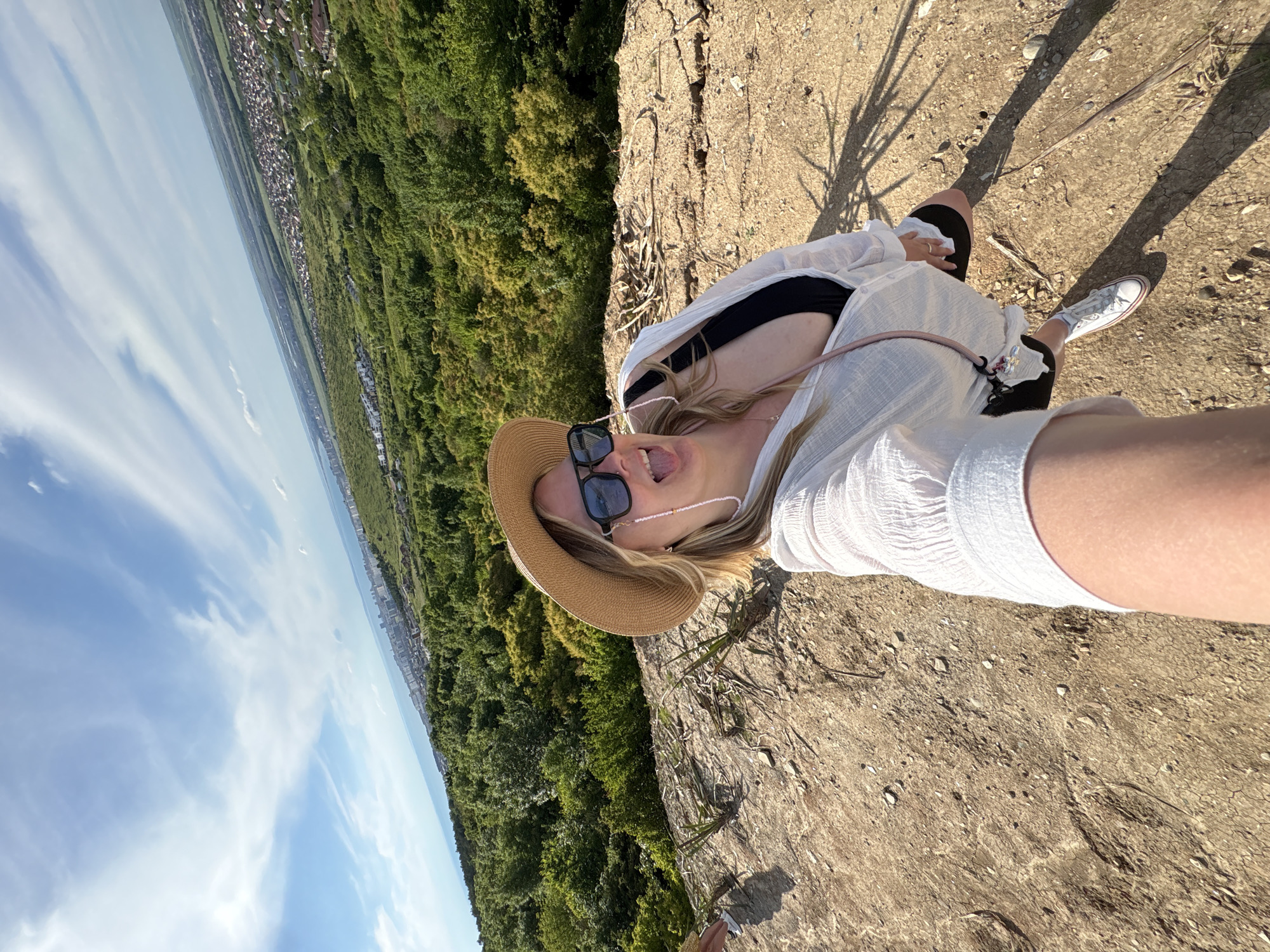 A young woman is captured in a selfie-style shot from a high vantage point, looking down at the camera with a playful expression, tongue slightly out. She has blonde hair, wears sunglasses, a light brown straw hat, a white semi-sheer long-sleeved top over a black top, and black shorts. White sneakers are visible on her feet.
The immediate foreground shows dry, light brown, rocky ground with sparse vegetation and long, distinct shadows, suggesting early morning or late afternoon sun. Below her, a vast landscape unfolds with dense green forest covering hillsides. In the mid-ground, a sprawling town or city with numerous buildings can be seen amidst the greenery. The horizon stretches into the distance, with a clear blue sky and scattered wispy white clouds overhead. The setting appears to be a natural overlook or cliff edge, offering a panoramic view of the surrounding area. There is no visible text in the image.