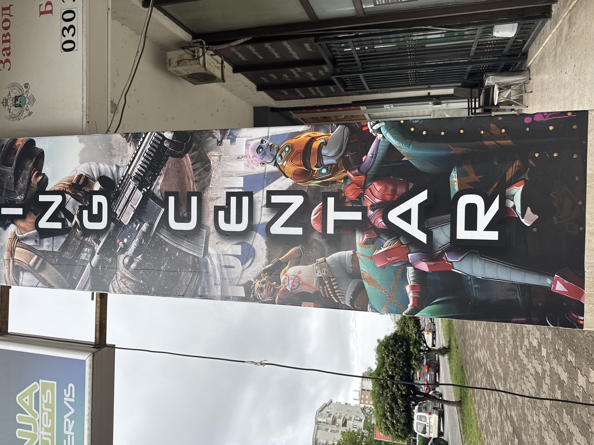 A sign reading "INUENTAR" in white capital letters is mounted in front of a storefront. The sign is decorated with images of video game characters. The background behind the storefront is a building with an air conditioning unit and a sign in Cyrillic script and numbers, possibly an address. A utility wire runs across the foreground. The street scene shows a tree-lined road with parked cars and apartment buildings in the distance. The image appears to be taken in Šušanj, Montenegro.
