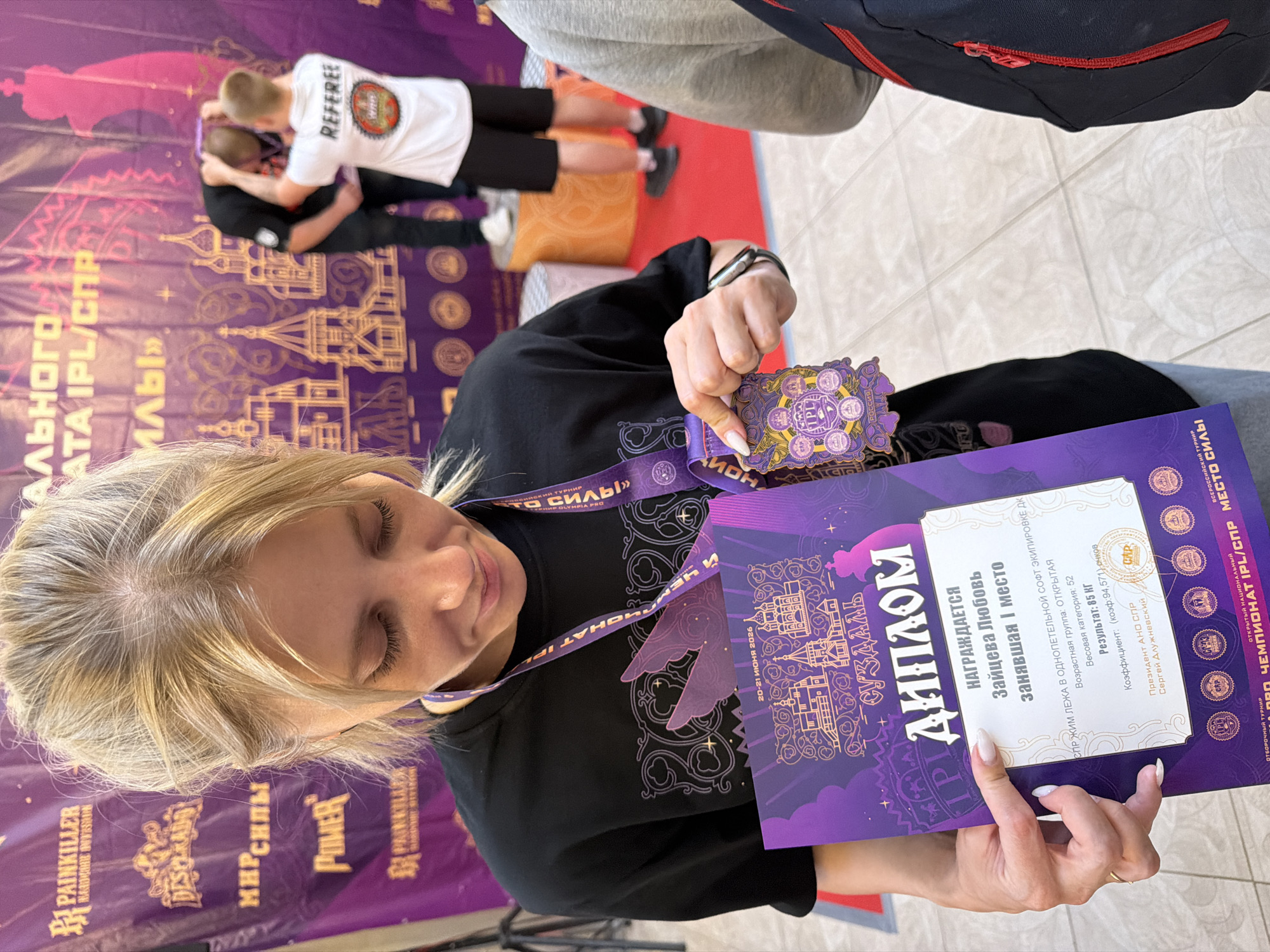 A young woman with blonde hair tied back holds a purple diploma and medal. She is wearing a black t-shirt and looking down at the diploma, which is partially obscured by her hand and the medal. The diploma features gold lettering and decorative elements, including depictions of buildings. The medal, also purple and gold, has several circular emblems. A purple lanyard with text is around her neck. In the background, a large purple banner displays text and images of ornate buildings. To the right, two men are visible on a stage. One man, wearing a white t-shirt with "REFEREE" printed on the back, is placing a medal around the neck of another man in a black shirt. The scene appears to be an awards ceremony or event.