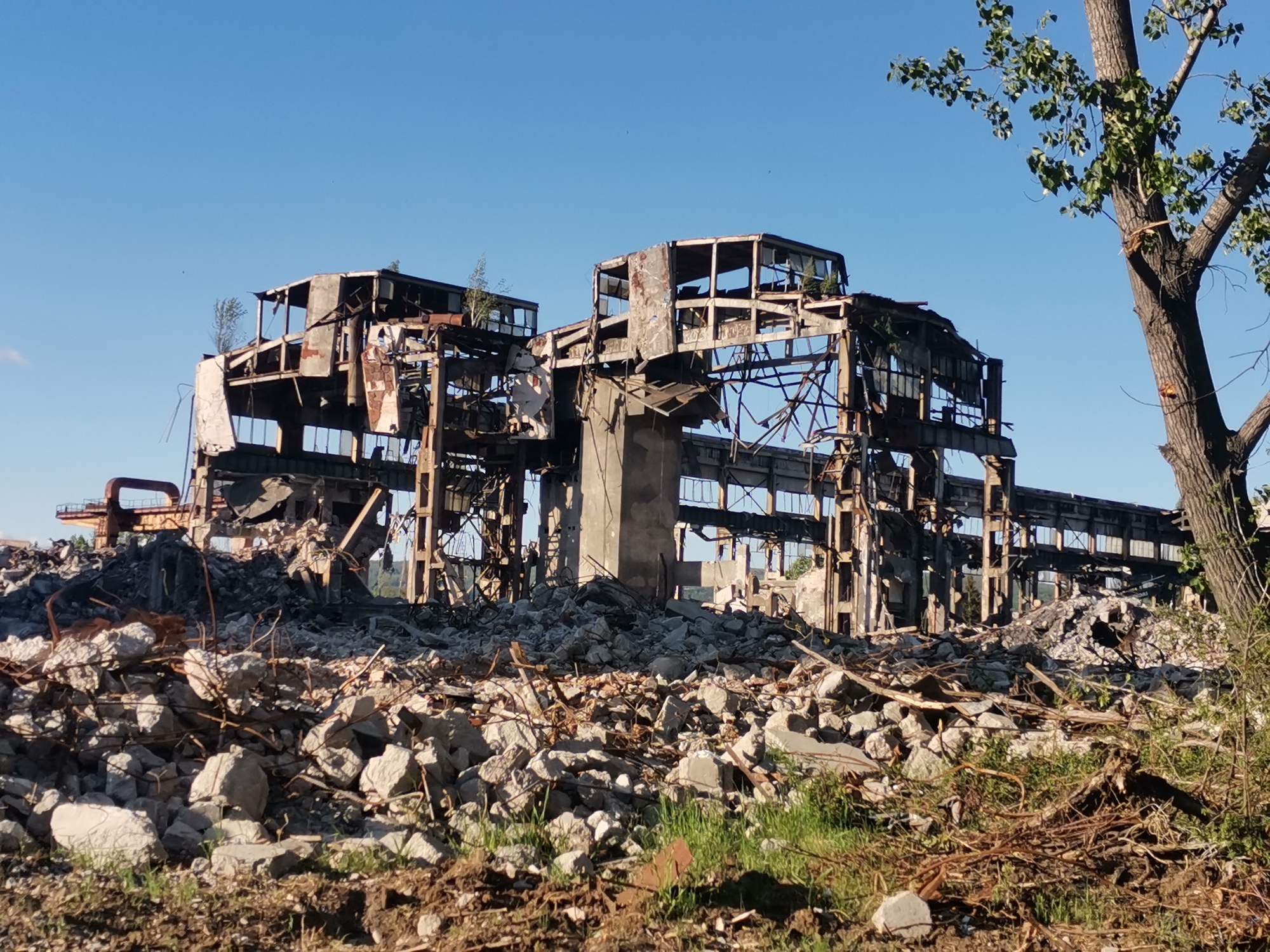 The image depicts a demolished industrial building in Zhivkovo, Bulgaria. The structure is heavily damaged, with exposed metal beams, concrete pillars, and debris scattered around. Rubble consisting of concrete, stone, and twisted metal covers the foreground. A tree with green leaves stands on the right side of the frame. The sky is clear and blue.