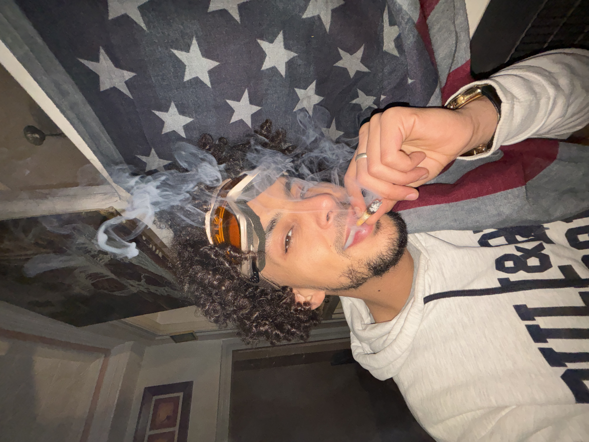 A young man with curly hair and tan skin is smoking. He is wearing goggles with orange lenses and a light gray hooded sweatshirt with a dark stripe and lettering. He has a gold watch on his left wrist and a ring on his left hand. Smoke emanates from the cigarette he holds in his right hand. He is lying on a blanket with a pattern of stars and stripes, possibly an American flag design. The background reveals a room with off-white walls, framed pictures, and ceiling fixtures. The photograph was taken from a low angle, looking up at the subject. There is no information to support the location being Tanger طنجة, Morocco.