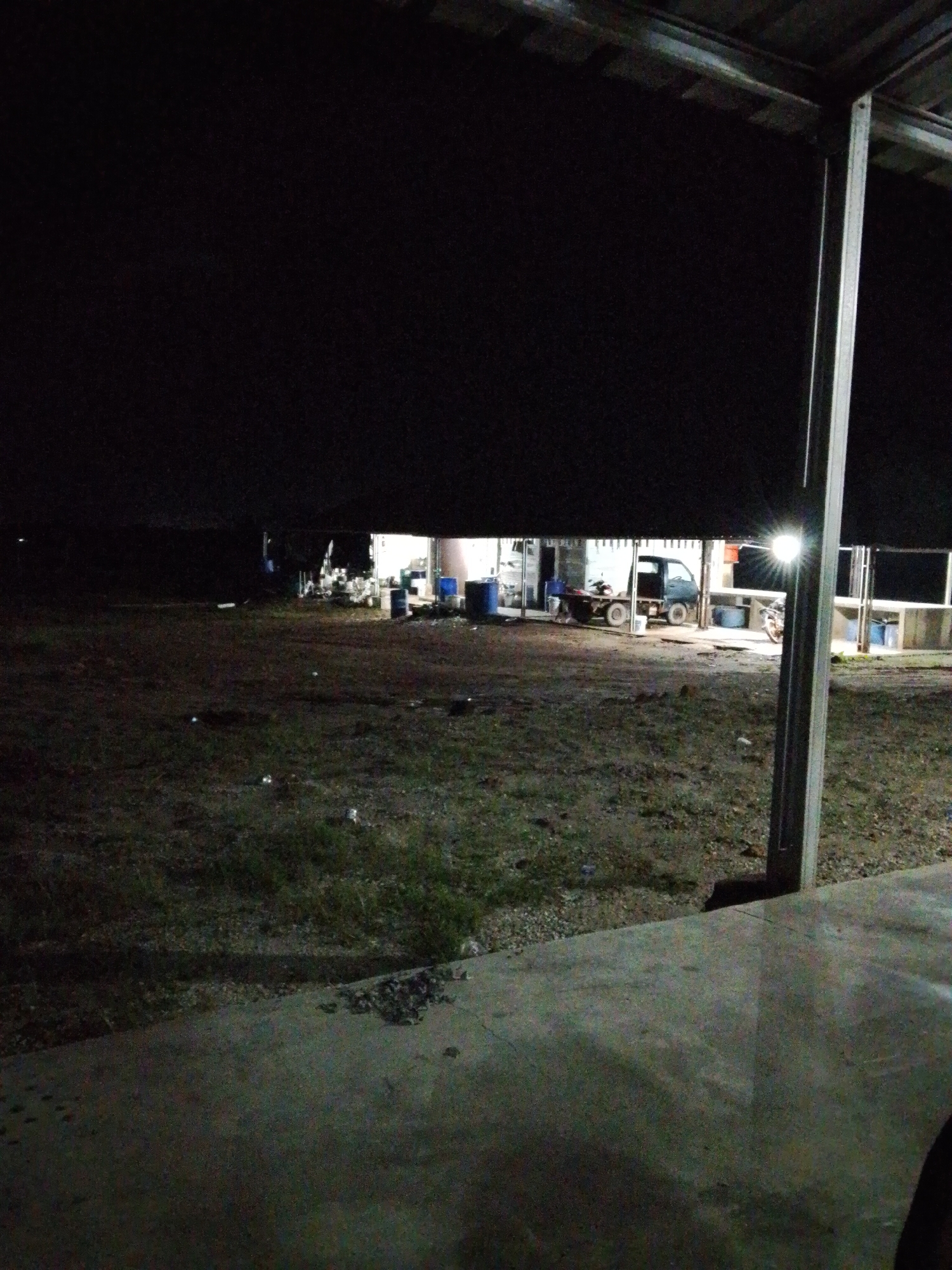 The image shows an outdoor scene at night, likely in Bukit Ketok, Indonesia. In the foreground, a concrete surface is visible. Beyond this, there's an area of unpaved ground with some vegetation. In the background, there is a brightly lit building with an open front. Inside or near the building are parked vehicles, including a truck and a motorcycle. Several large barrels are also present near the building. A metal support beam is located on the right side of the image. The overall environment is dark.