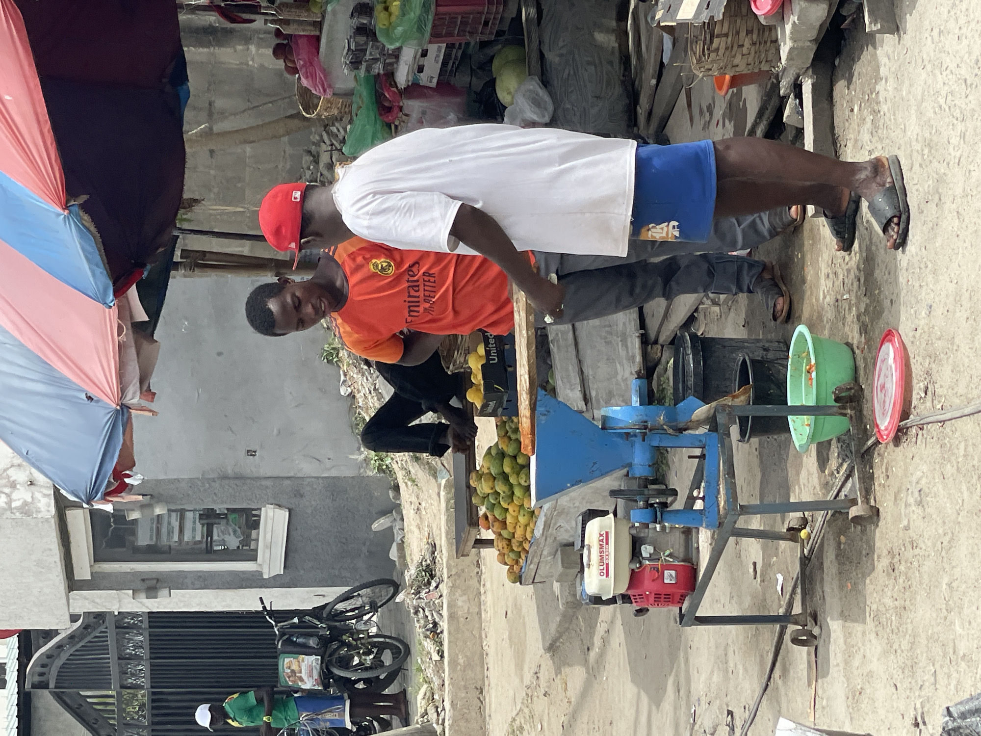 The image shows two men at a roadside fruit stall in what appears to be Lekki, Nigeria. One man, wearing a red baseball cap and an orange soccer jersey, is operating a fruit-processing machine, likely for juicing. The other man, in a white t-shirt and blue shorts, is standing nearby. The fruit being processed appears to be mangoes, a large quantity of which is stacked on the stall counter. The machine is a blue metal structure with a red and white engine attached. There are various buckets and containers around the machine, presumably for holding the processed fruit. The background shows a street scene with a building, an umbrella shading the stall, and a parked bicycle.