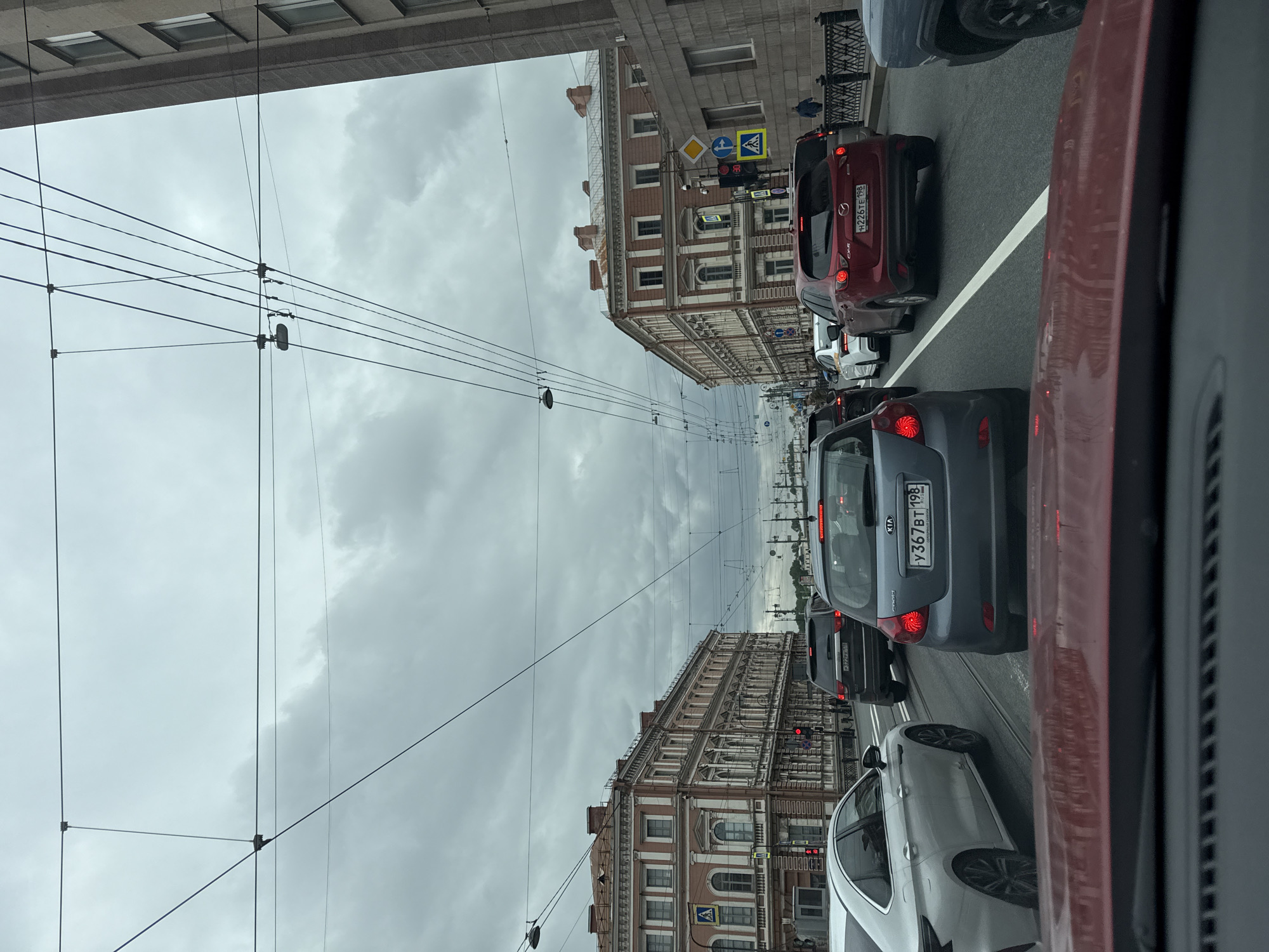 The image depicts a street scene in an urban environment, characterized by a traffic jam. The location appears to be in Saint Petersburg, Russia, based on the architectural style of the buildings and the license plates of the vehicles. The sky is overcast, suggesting a cloudy day. Several cars are visible, with a gray Kia in the center. There are overhead tram lines. The buildings on either side of the street are of a classical design. Pedestrian crossings are indicated by street signs. The photograph was taken from inside a vehicle, looking out onto the street.