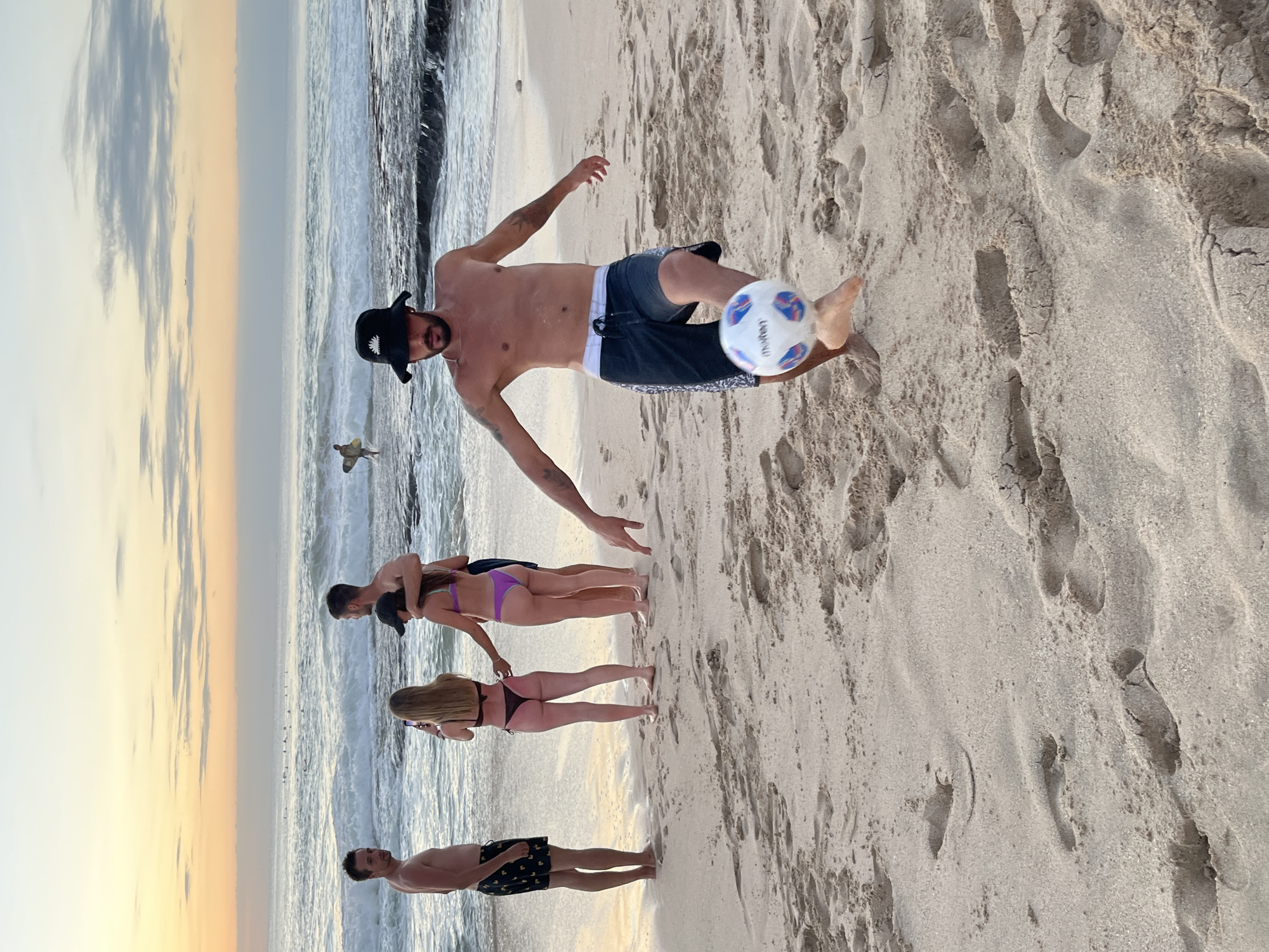 In Kuta Selatan, Indonesia, on a sandy beach at sunset, a group of people are gathered near the ocean. The sky displays soft hues of orange and yellow. In the foreground, a shirtless man wearing a black bucket hat and dark patterned swim trunks is playing soccer, kicking a white and blue ball. To his left, further down the beach, a man in dark patterned shorts stands looking at the camera. Behind him, three people are standing together: a woman in a dark bikini taking a picture with her phone, and a man with his arm around a woman wearing a purple bikini. In the background, waves roll onto the shore, and several surfers are visible in the distance, some with their surfboards. The sand is marked with footprints.