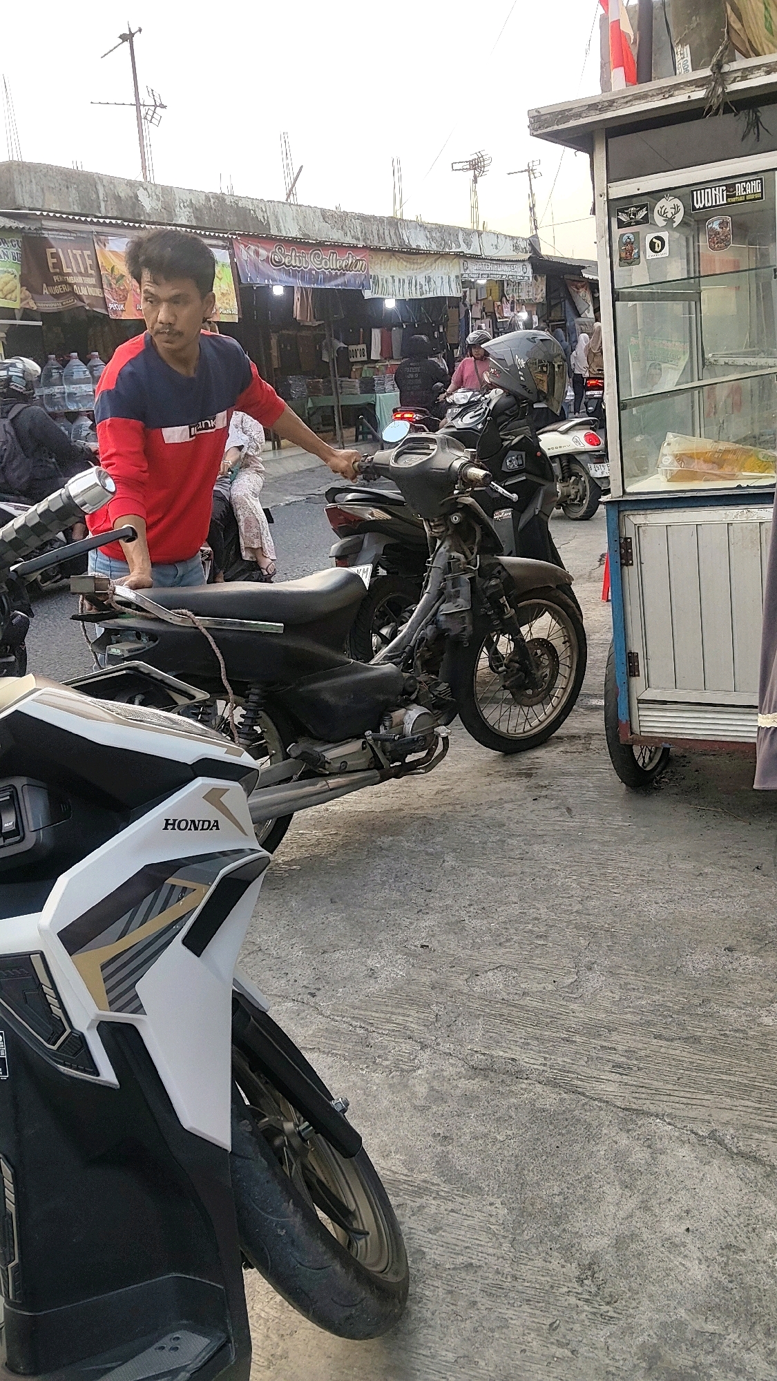 The image depicts a street scene in Mekarmukti, Indonesia, likely near a market or small shops. In the foreground, a person wearing a red and blue long-sleeved shirt and denim jeans is standing next to a black motorcycle. The person is holding onto the handlebars and appears to be looking to the right. To the right of this motorcycle, a white and black Honda scooter is partially visible. Behind the first motorcycle, another black motorcycle with a rider wearing a helmet is parked. Further in the background, there are several market stalls with banners, including one that reads "Elite" and another that says "Selvi Collection." A food cart or small kiosk with glass display cases and a sign that reads "WONG OCANG" is also present on the right side of the image. The ground is paved with concrete. The sky is overcast, suggesting it may be late afternoon or early morning.