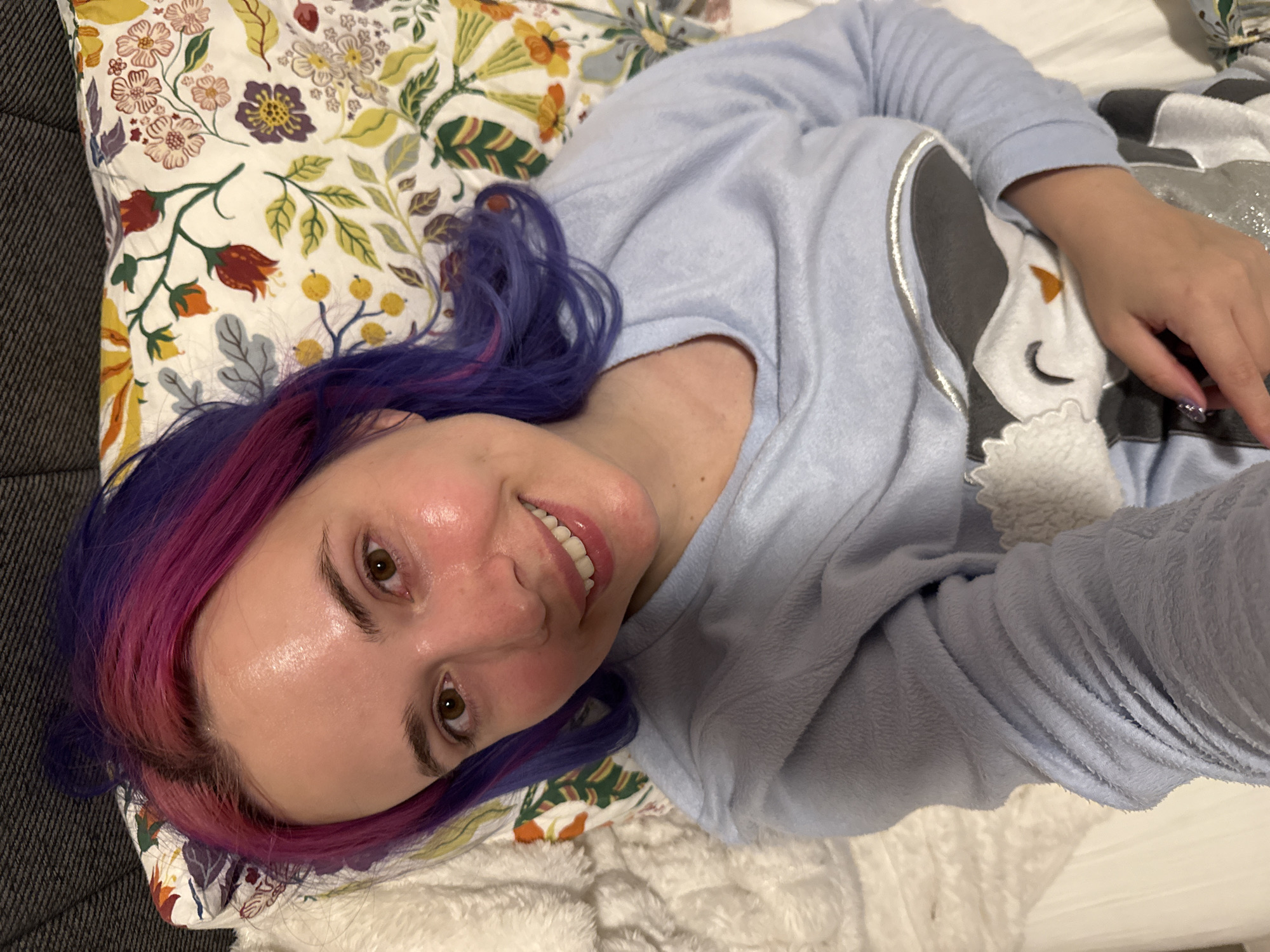 A woman with dyed purple and pink hair lies on a bed. Her head rests on a patterned pillow, and she wears a light blue, long-sleeved shirt and pajamas with an animal design. Her right hand rests on the pajamas. The location is not discernible from the image.