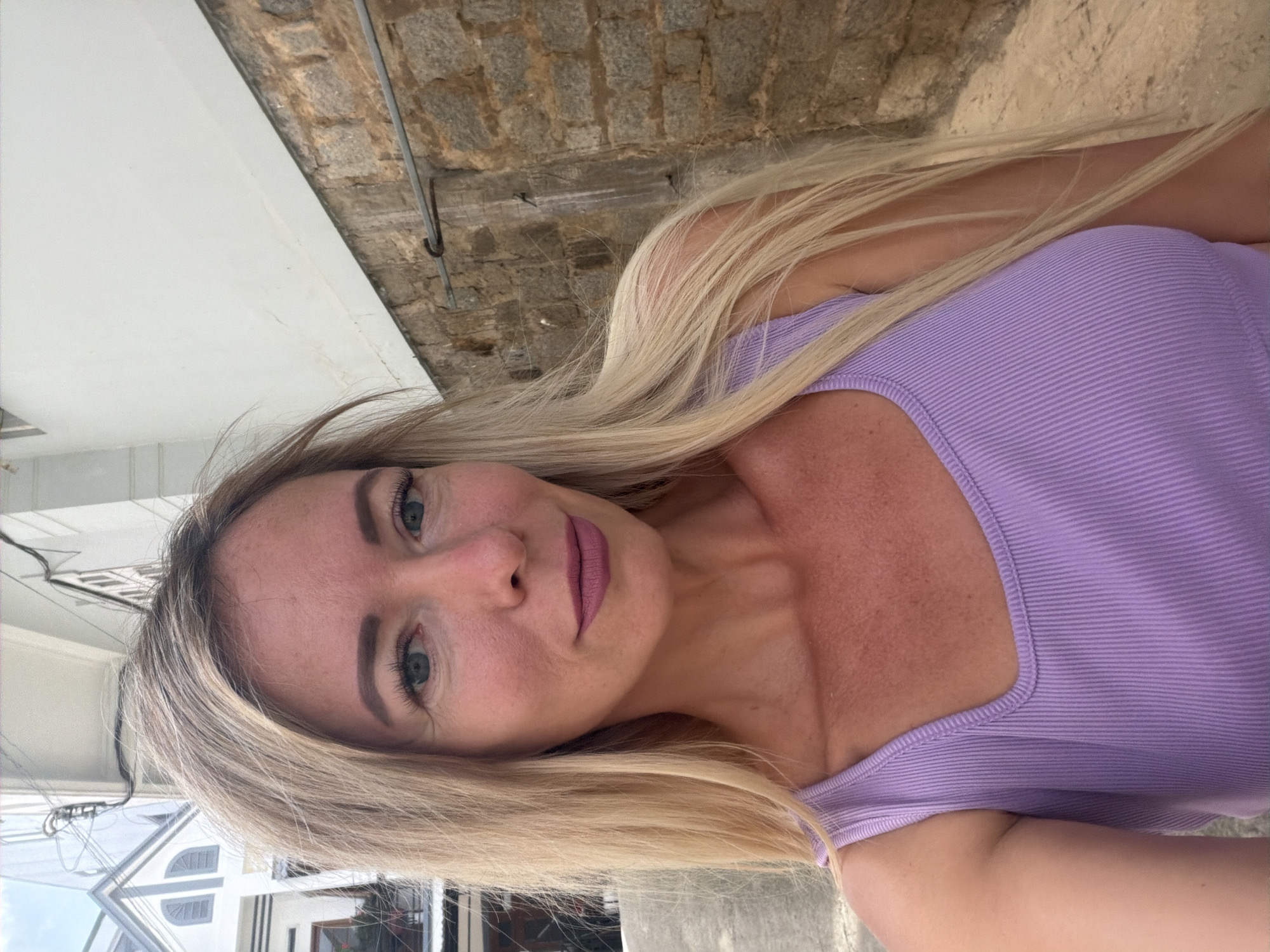 A fair-skinned woman with long blonde hair and blue eyes is standing outdoors in a narrow alleyway in Da Lat, Vietnam. She is wearing a light purple ribbed tank top. The background consists of a white building with architectural details, a stone wall, and electrical wires. The lighting suggests it is daytime.