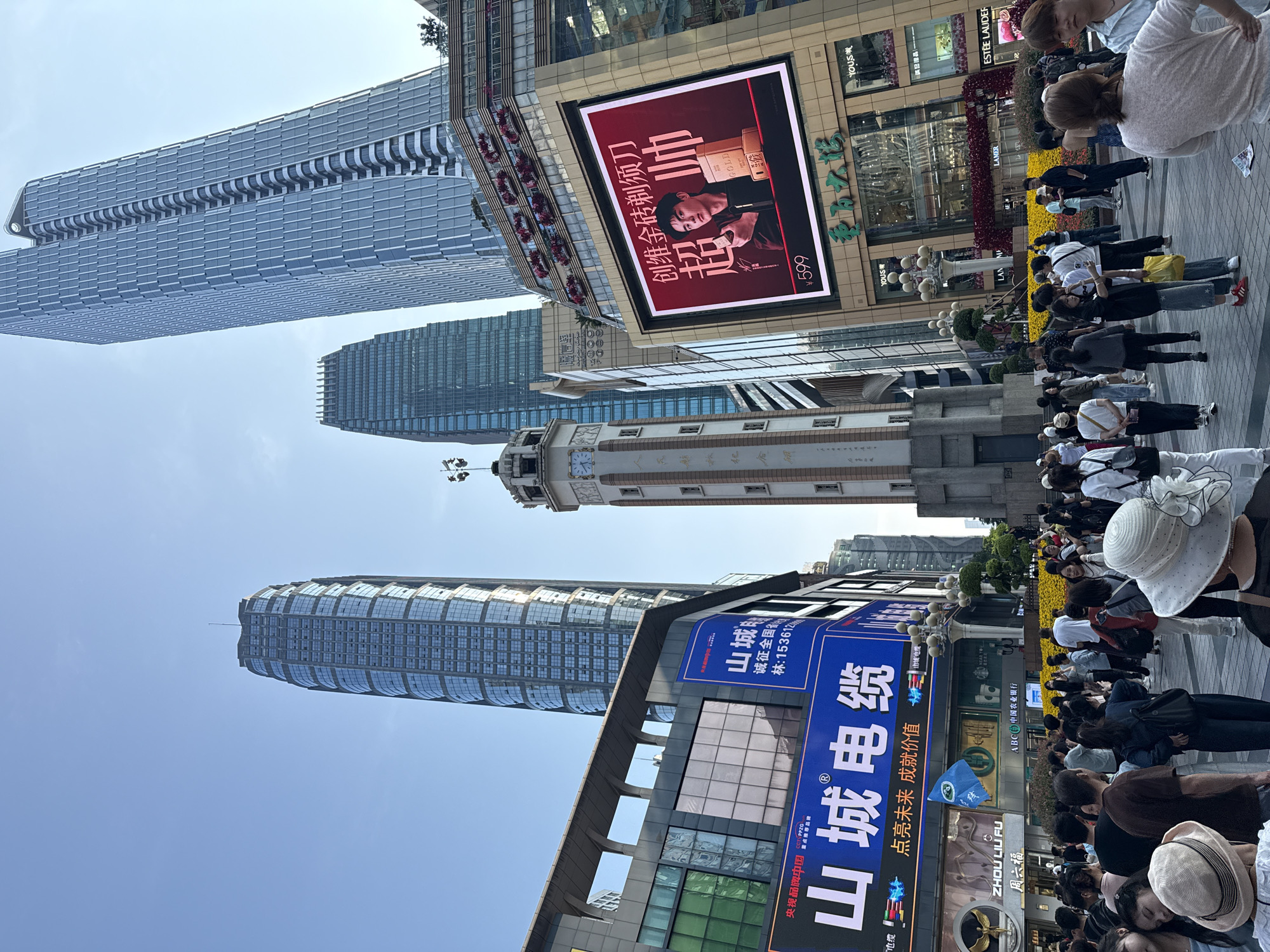 The image depicts a street scene in the Yuzhong District, China, characterized by high-rise buildings and a crowd of people. Prominent structures include modern skyscrapers with glass facades and a clock tower. Large electronic billboards display advertisements, including one for "Shan Cheng Dian Lan" (Mountain City Cables) and another for a "Chuangwei Jinzhuan Tixudao" (Skyworth Gold Brick Razor). People are visible walking and standing in the street, with various clothing styles and accessories. Some wear hats, and others carry bags. The environment includes storefronts with visible brand names like "YOUS," "ESTEE LAUDER," "LAN," and "LAMER." A flower arrangement is also visible along the pedestrian area. The sky is clear and blue, suggesting a bright, sunny day.
