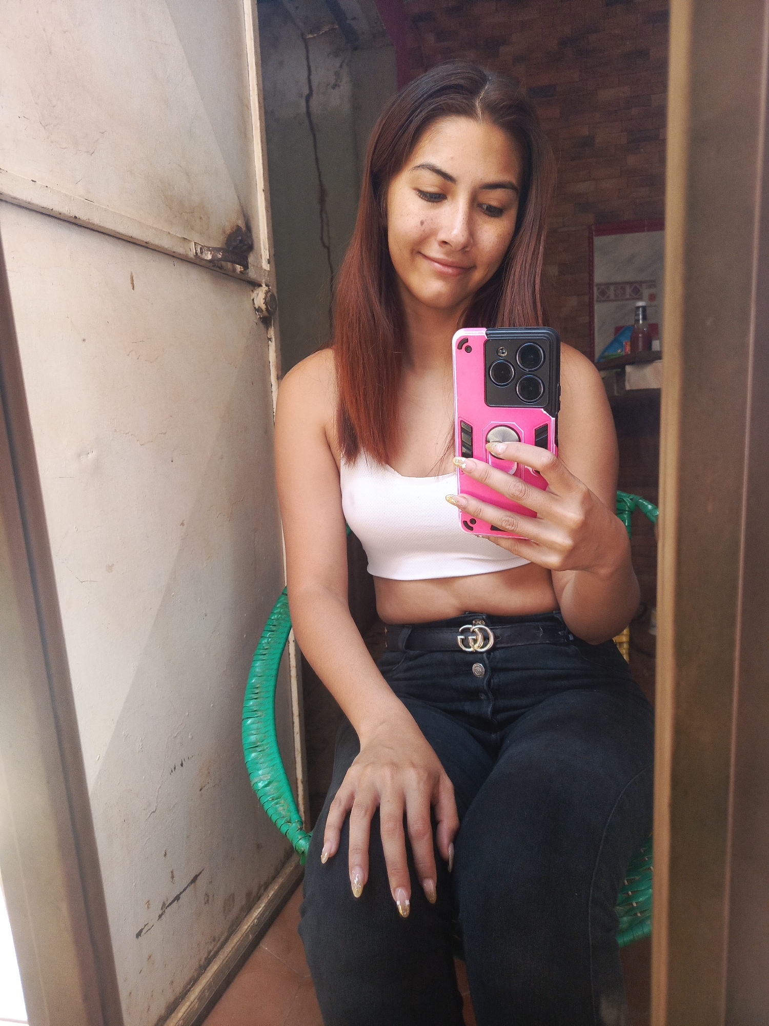 A woman sits in a green chair in what appears to be an indoor/outdoor space. The wall to her left is light-colored and worn. She is fair-skinned with brown hair, wearing a white crop top and dark jeans with a Gucci belt. She is holding a pink-cased smartphone in her right hand, looking at it and taking a selfie. Behind her is a brick wall and some shelves. The scene is a reflection in a mirror in the city of Juan Griego, Venezuela.