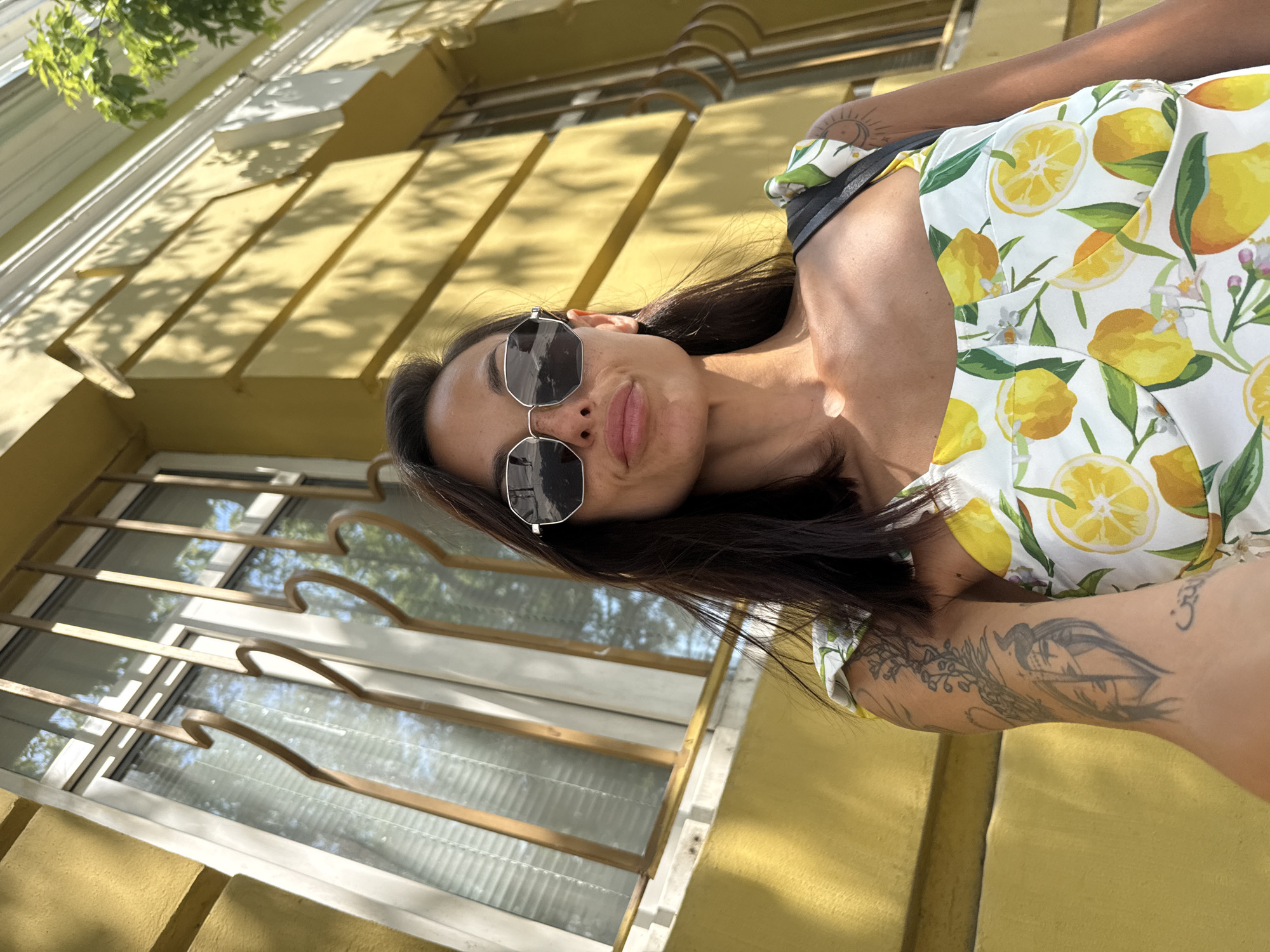 A young woman with dark hair, wearing sunglasses and a white dress with a lemon print, takes a selfie in front of a yellow building. The building has windows with decorative iron bars and architectural detailing. The woman's left arm is visible, showing tattoos. The scene suggests an outdoor location on a sunny day, possibly in the city of Pleven, Bulgaria.
