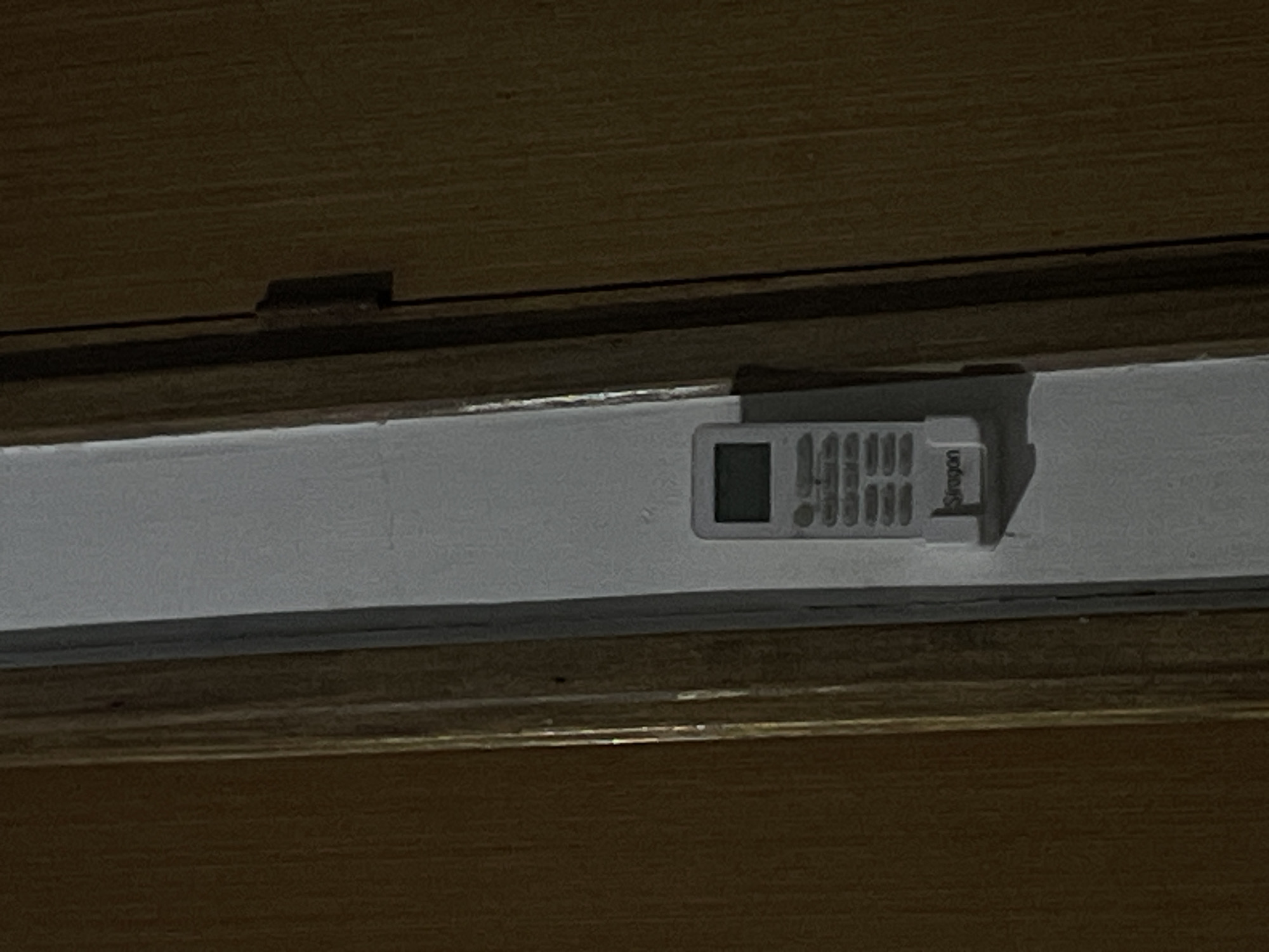 This is a medium shot photograph of a small, white device attached to a wooden surface. The device is rectangular with rounded corners and appears to be a digital thermometer or sensor. It has a small screen displaying a few digits, and below the screen are several small buttons. The brand name "Oregon" is visible on the right side of the device, printed in black. The device is mounted on a white strip, which is itself attached to a wooden panel. The lighting is dim, casting shadows and highlighting the texture of the wood grain. The scene appears to be indoors, possibly a home or office setting, and there are no people or other objects clearly visible. The focus is entirely on the small white device and its immediate surroundings.