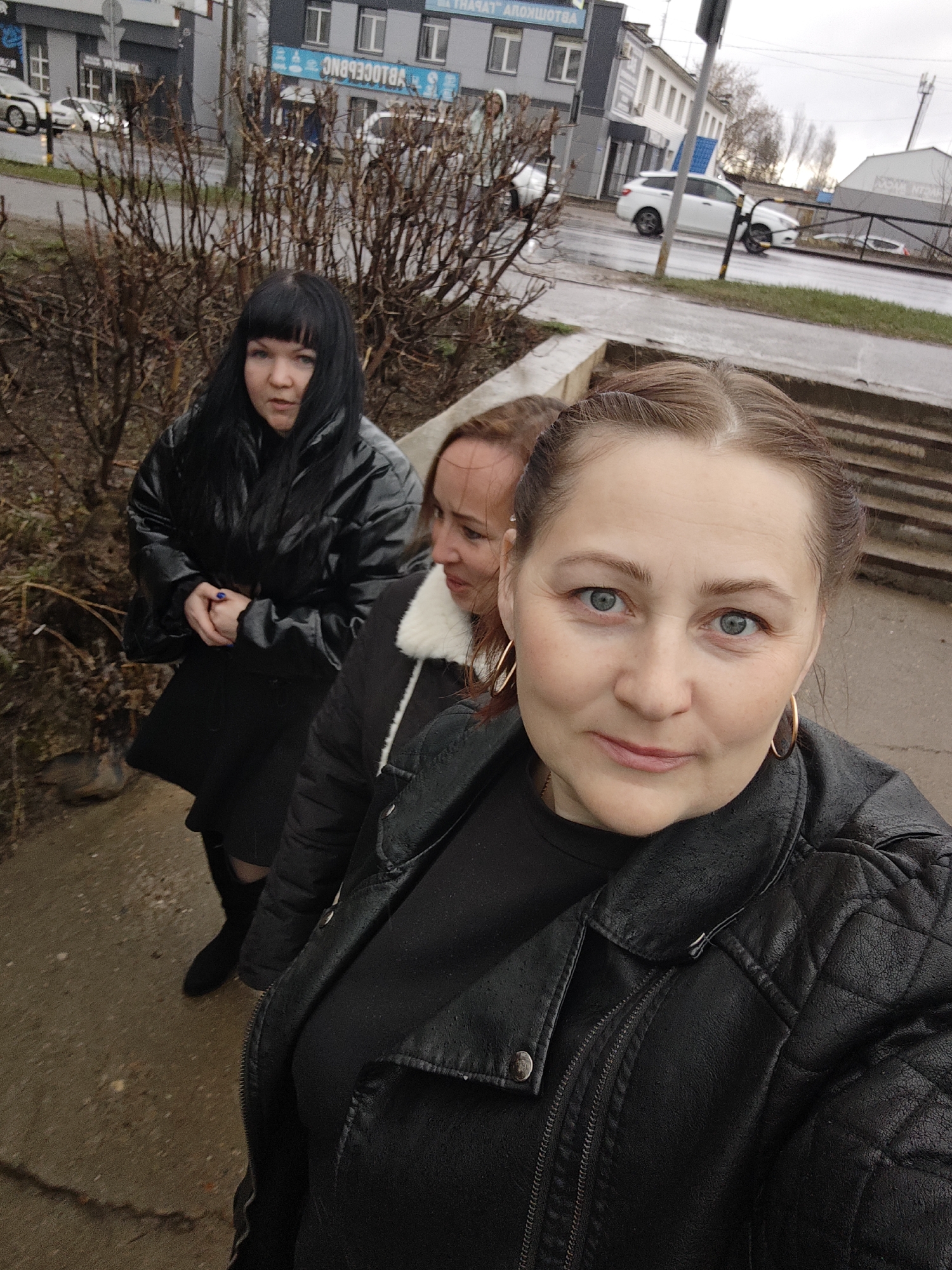 Three women are standing on a sidewalk in what appears to be an urban environment. The woman in the foreground is holding the camera; she has light skin, fair hair pulled back, blue eyes, and is wearing a black leather jacket and large gold hoop earrings. Behind her and to the left, the second woman has dark hair with bangs, pale skin, and is wearing a shiny black coat and dress. The third woman is standing between the first two, partially obscured. She has light skin and brown hair. The women are all facing the camera. They are standing next to a bare bush and a low concrete wall. Behind them, a street scene includes cars, buildings, and a sign in Russian. The surroundings suggest the location is in a city, likely Chaikovsky, Russia, based on the context. The sidewalk appears wet.