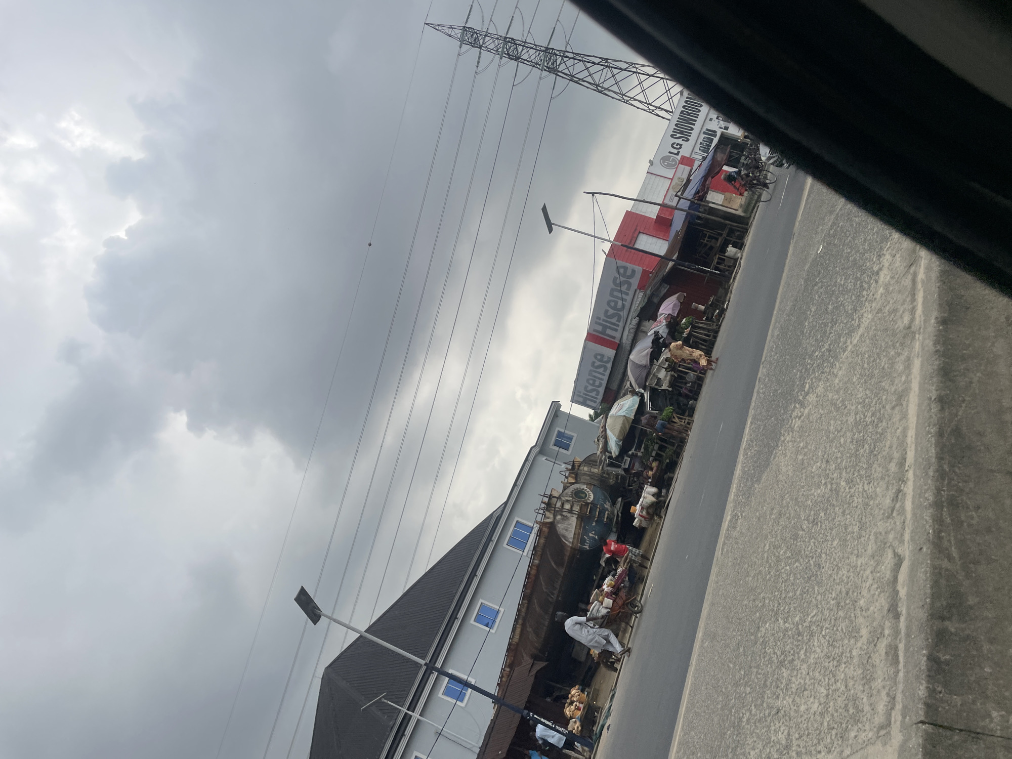 This image captures a vibrant street scene in Eliozu, Nigeria, under a heavily overcast sky. The photograph is taken from a slightly elevated and tilted perspective, possibly from a vehicle.
The sky is dominated by thick, grey clouds, suggesting either impending rain or a generally cloudy day. A prominent feature in the upper left is a large metal electricity pylon or transmission tower, with numerous power lines stretching horizontally across the frame.
Along the right side of the road, a series of commercial buildings line the street. One building clearly displays a large red and white "Hisense" sign, with "LG SHOWROOM" visible above it, indicating a retail presence for electronics brands. Adjacent to it, another building features light grey walls, a dark grey gabled roof, and multiple blue-tinted windows. In front of these structures, a bustling market scene is in progress, with various goods displayed under umbrellas and makeshift awnings. Several people are visible, engaged in activities such as walking, standing by stalls, or pushing carts. One individual in light-colored clothing is pushing a cart laden with items. A large, dark blue, circular object resembling an industrial tank or container is noticeable on a flatbed structure near the central building.
The road, made of asphalt, runs parallel to the buildings. A concrete curb separates the main road from a dirt or gravel shoulder on the far right. Modern street lights with rectangular fixtures are installed along the roadside. Bicycles are also visible, parked or being ridden, contributing to the street activity. The overall impression is one of active commerce and everyday life in a bustling urban or semi-urban area.