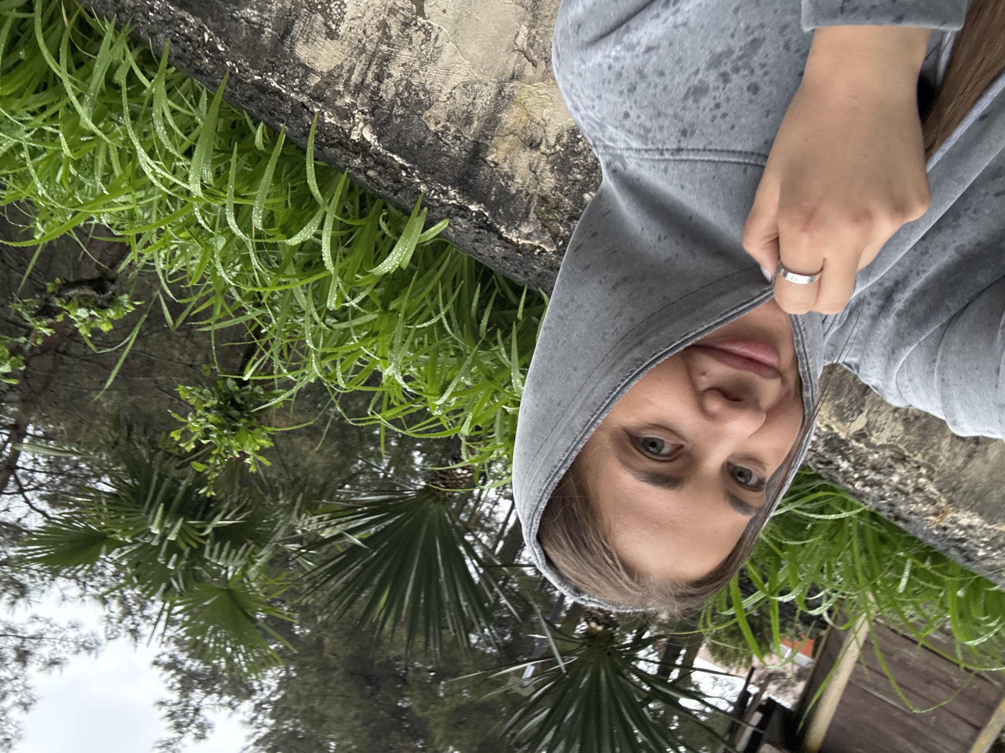 A young woman stands in front of lush green foliage and palm trees in Sosnovy, Russia. She is wearing a gray hoodie and holding the hood up to partially cover her face. She is wearing a ring on her finger. A concrete structure is visible near the green foliage. The sky appears overcast.