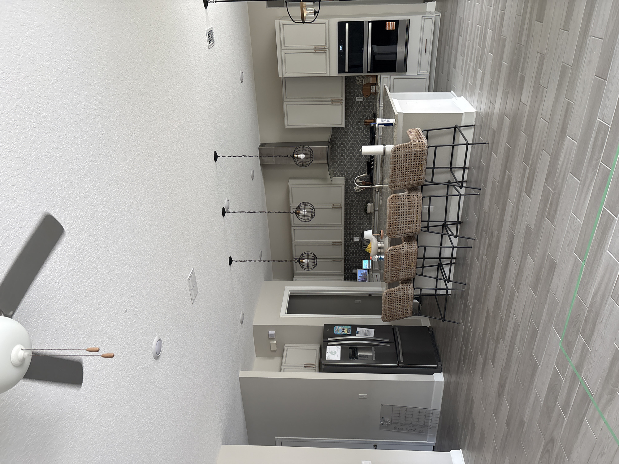 The image depicts an interior scene of a modern kitchen and dining area. The flooring is a light gray tile laid in a wood-plank pattern. In the foreground, a kitchen island with a light-colored countertop is visible, featuring three woven bar stools with black metal legs. Behind the island, a stainless steel refrigerator is situated to the left, and a double oven unit is to the right. The kitchen cabinets are a light gray with minimalist hardware. The backsplash behind the stove features a gray hexagonal tile pattern. Above the island, three industrial-style pendant lights with black wire cages are suspended from the ceiling. A ceiling fan with two wooden pull chains is also visible. The walls are painted a neutral light gray. The scene appears to be a residential kitchen located in Bradenton, Florida, United States.
