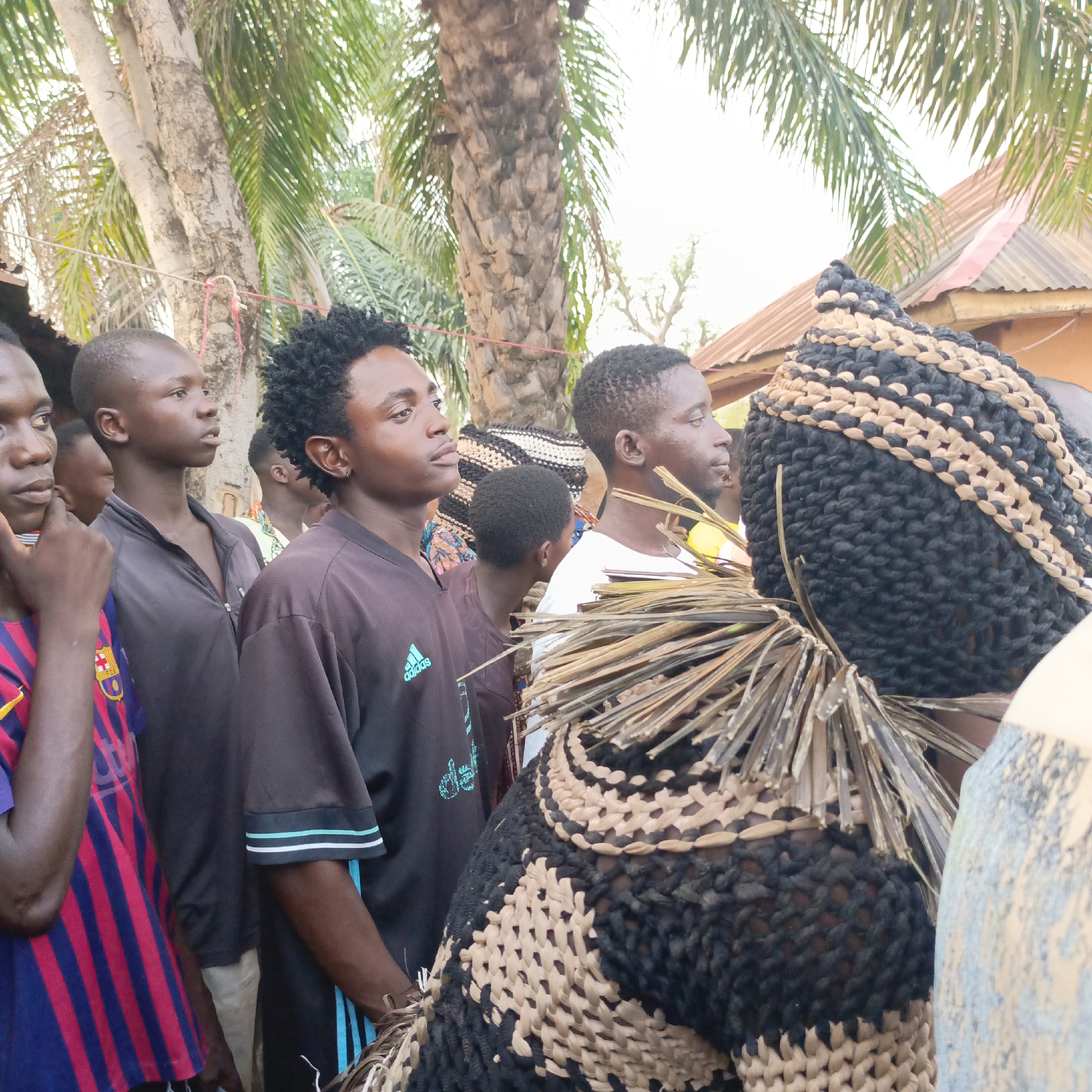 A photograph taken in Akwanga, Nigeria, shows a group of young men gathered around an individual wearing a traditional woven mask and costume. The mask is crafted from black and beige woven material, with a fringe of dried straw-like elements around the neck area. The person wearing the mask is positioned towards the right side of the frame. To the left, there are at least five young men, most of whom are looking towards the person in the mask. They are wearing casual clothing. The background includes trees, including palms, and the corner of a building with a corrugated metal roof. The scene suggests a cultural event or performance.