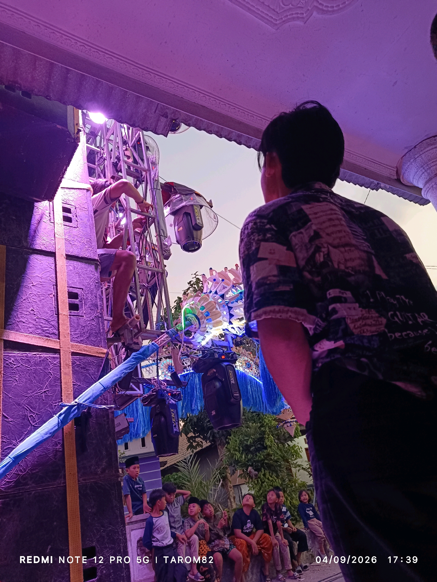 In Rejoagung, Indonesia, an outdoor event is underway, illuminated by purple stage lighting. A man, seen from behind, wearing a dark patterned shirt and dark pants, stands in the foreground, facing away from the camera. He appears to be watching the activities at the event.
In the midground, another man, wearing a white t-shirt and shorts, is perched precariously on a tall ladder, which is attached to a large stack of black speakers. He is reaching out to adjust some equipment on a metal truss structure. This truss is adorned with colorful, flower-like decorations, possibly part of a stage setup. Several professional-grade stage lights are mounted on the truss. Below the truss and speakers, a group of children, dressed casually and some wearing skullcaps, are seated on a low wall, looking up at the spectacle. They appear engaged and curious.
The overall environment suggests a festive gathering, possibly a religious celebration or a community event, given the attire of some of the children and the presence of stage equipment. The date stamp on the image indicates "04/09/2026 17:39", suggesting the photo was taken on September 4, 2026, at 5:39 PM. The text overlay "REDMI NOTE 12 PRO 5G | TAROM82" likely refers to the device used to capture the image and possibly a username or location tag.