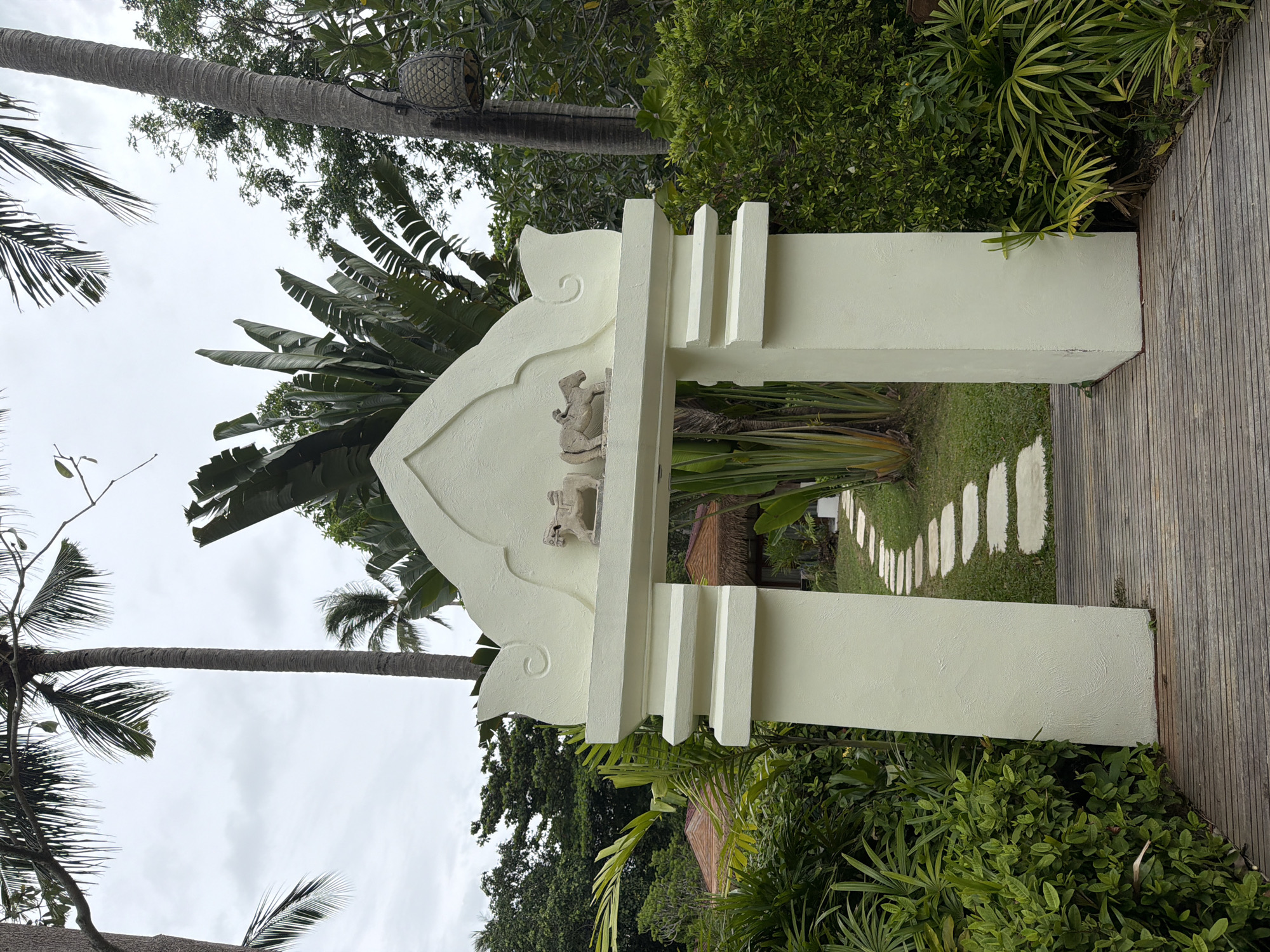 In the Ko Samui District of Thailand, a decorative archway stands on a wooden walkway, framing a stone pathway that winds through lush greenery. The archway is painted a pale cream color and features two small, light-colored sculptures of figures on horseback at its apex. Flanking the entrance, dense tropical foliage creates a vibrant green backdrop. Beyond the arch, a path of irregularly shaped stone pavers leads towards the blurred shapes of thatched-roof buildings. Tall palm trees and large-leafed plants are visible, contributing to the tropical atmosphere. The sky above is overcast, suggesting a diffused light condition.