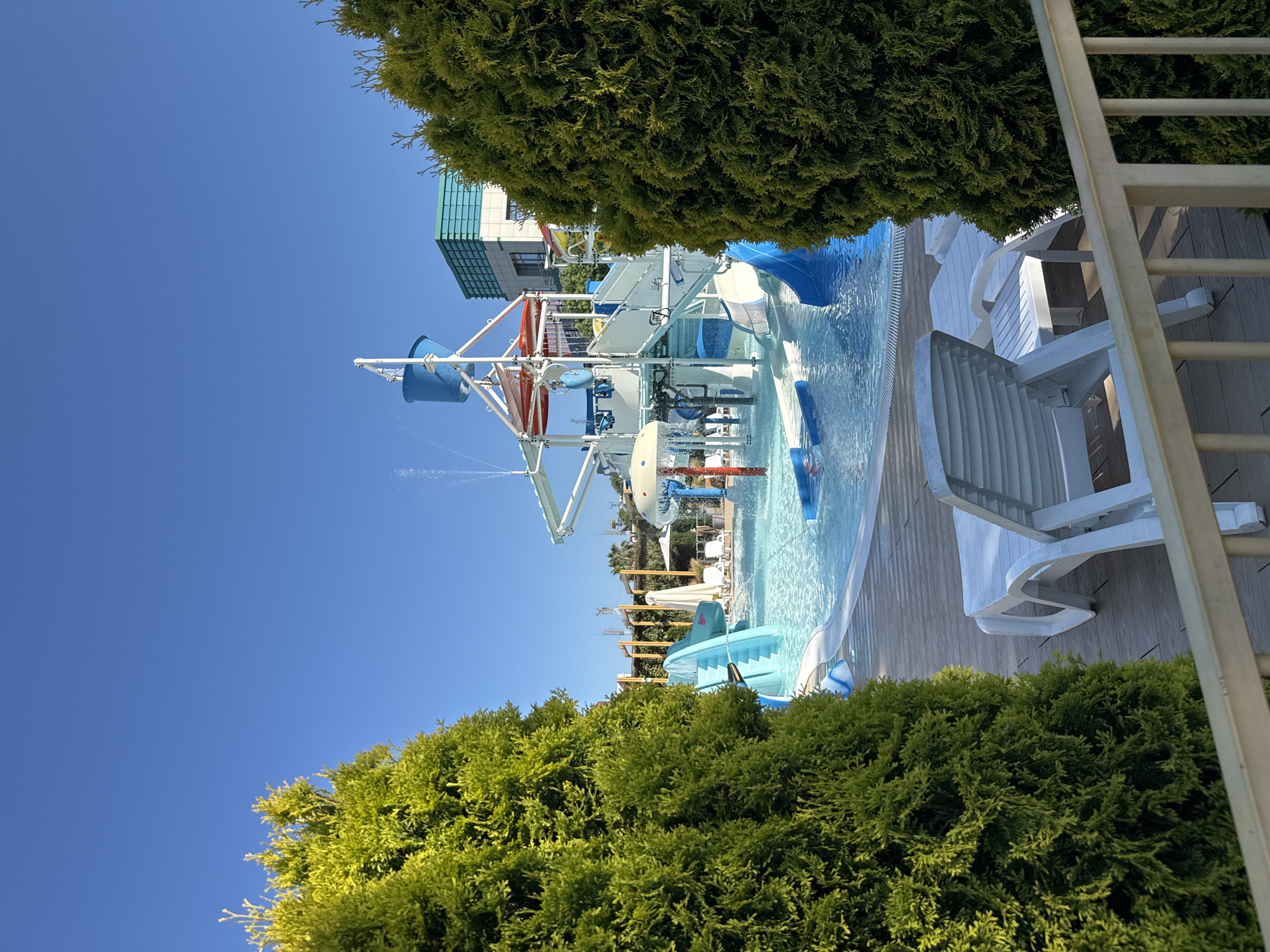 The image depicts a water park in Sochi, Russia, bathed in bright sunlight under a clear blue sky. In the foreground, two white plastic lounge chairs are positioned on a wooden deck. To the left and right, dense green coniferous trees frame the view, with their branches obscuring parts of the scene.
The central focus is a colorful water park structure with slides, platforms, and water features. A large blue bucket at the top of the structure appears to be tipping water. Several blue and white slides descend into the clear blue pool. Water jets are spraying in various areas, creating a lively atmosphere. In the background, there is a modern building with a light-colored facade and turquoise accents, likely part of the resort. Beyond that, hints of other structures and vegetation can be seen, suggesting a sprawling resort complex. The water in the pool is calm and inviting, reflecting the blue sky and the surrounding structures. The overall impression is one of a vibrant and fun recreational area on a sunny day.