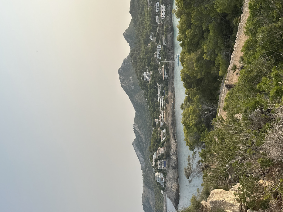 The image presents a panoramic coastal scene located in Capdepera, Spain. A hazy, light-gray sky dominates the upper portion. In the middle ground, a tranquil bay with light blue-green water extends, bordered by a rocky coastline. Numerous white and light-colored residential buildings, predominantly houses, are scattered across the green, tree-covered hillsides surrounding the bay. One white sailboat is anchored in the bay's center-right section. The foreground is densely covered with green shrubs and trees, with a visible unpaved path bordered by a low stone wall winding through it from left to right. A large, light-colored rock formation is situated in the lower-left foreground. Undulating, rugged mountains, also covered in vegetation, form the background. No individuals or direct actions are discernible within the frame.