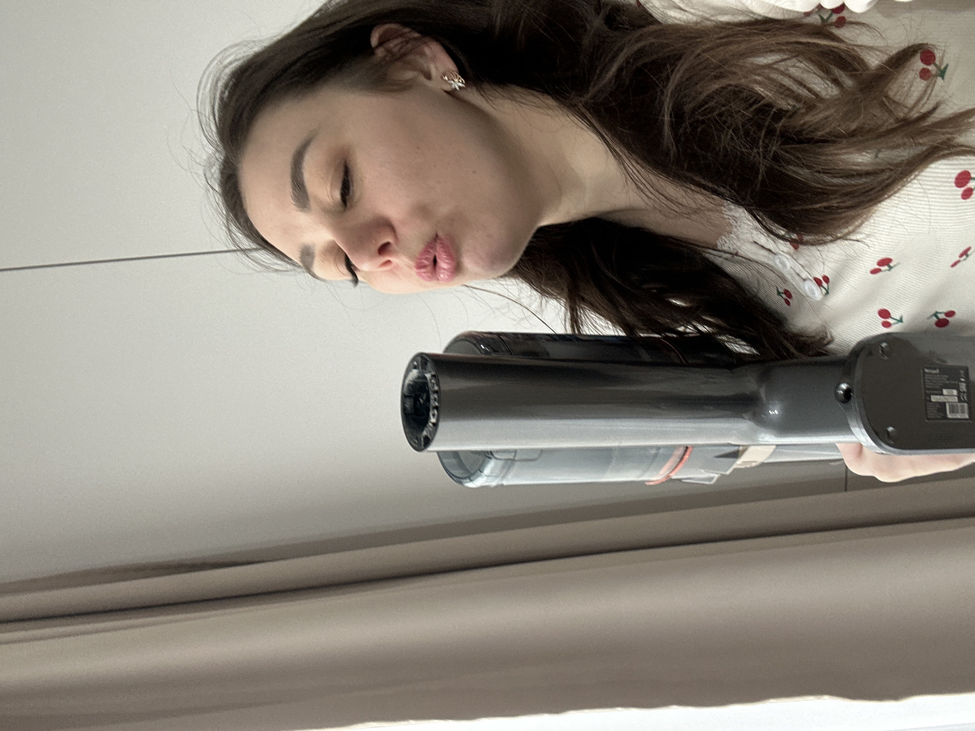 The image is oriented on its side, showing a young woman from the chest up, engaged in a playful interaction with a handheld device. The woman, with long, wavy, dark brown hair, is positioned on the left side of the frame. Her head is tilted, and her eyes are partially closed as she pouts her lips in a "kissy face" or "duck face" expression, suggesting a lighthearted or humorous mood. She wears a light-colored top patterned with red cherries and features a white, possibly ruffled, collar. A small, decorative earring is visible in her visible ear.
Her right hand extends into the frame from the bottom right, holding a sleek, metallic grey and black handheld device. The device, which resembles either a portable hair styling tool or a small dustbuster, is pointed towards her face, with its nozzle or barrel clearly visible.
The background consists of a plain, light grey or off-white wall or ceiling, featuring a subtle horizontal line. Towards the bottom of the frame, a horizontal band of a darker, brownish-grey material, possibly a curtain or window frame, is visible. The lighting is bright and even, indicating an indoor setting. On the side of the handheld device, near her hand, a small, blurry label with unreadable text is present. There are no specific visual cues within the image to confirm the provided location context of Chaikovsky, Russia.