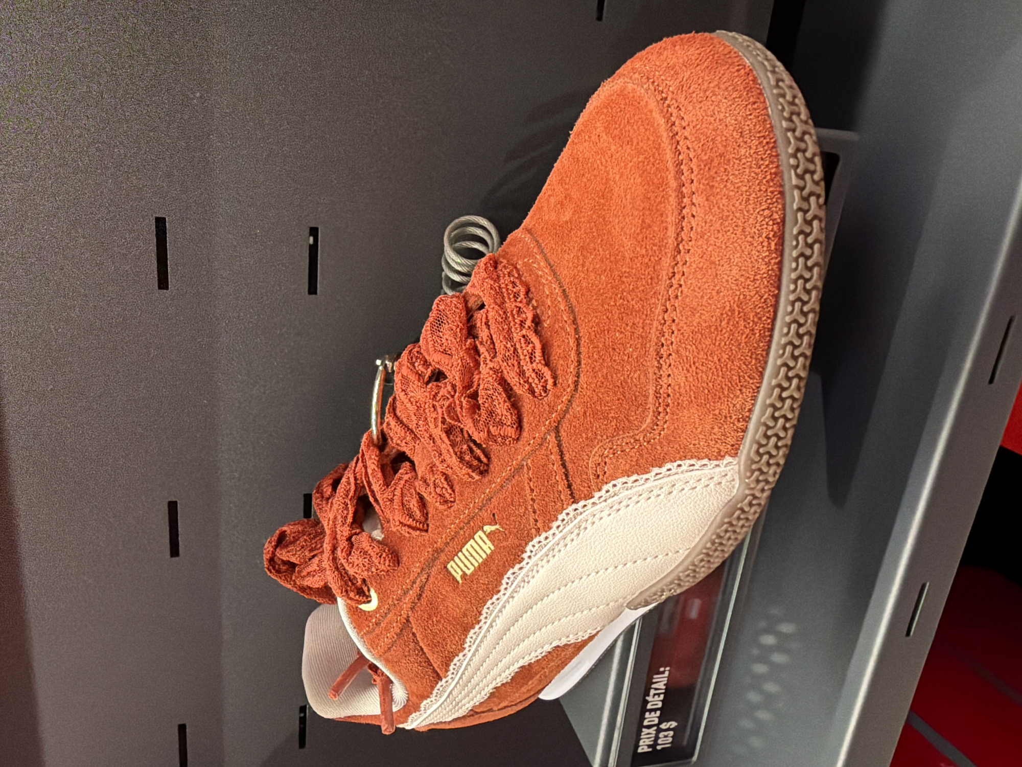 A close-up, eye-level shot shows a single, orange Puma athletic shoe displayed on a gray shelf. The shoe is positioned at an angle, facing right. It features a suede upper with a creamy white stripe detail along the side, trimmed with a delicate lace-like pattern. The laces are a matching orange hue and appear textured. The Puma logo is visible in gold on the side of the shoe. A security cable is coiled around the top of the shoe. In the lower left corner, a black price tag is partially visible with white text that reads "PRIX DE DÉTAIL: 103 $". The background consists of a dark, textured wall with horizontal slots. The shelf is dark gray and has subtle indentations along its front edge. The lighting is neutral, casting soft shadows. The scene appears to be in a retail environment, likely a shoe store.