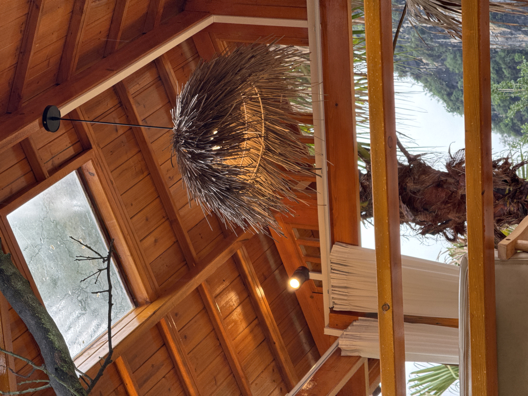 The scene is an indoor space with a wooden ceiling and walls, possibly a gazebo or a treehouse-like structure, possibly located in Guilin, China.
A circular, straw-like chandelier hangs from the ceiling via a black metal rod, positioned near a rectangular window. Outside the window, a tree branch is visible. A beam of light comes from a spotlight near the right side of the frame. Through an opening, foliage and a blurry landscape can be seen. Wooden planks are visible in the foreground, possibly part of a supporting structure. Blinds are partially drawn.