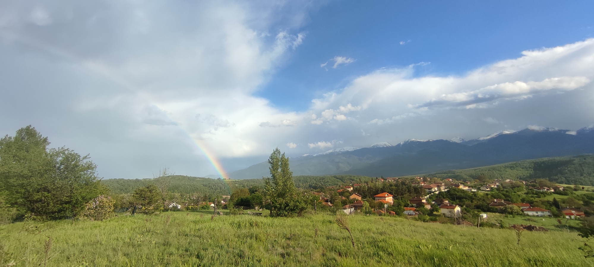 The image shows a vibrant rural scene in Gutsal, Bulgaria. A rainbow arches across the sky, its ends seemingly touching down in the green landscape. The foreground is dominated by a grassy field, dotted with occasional trees and shrubs. In the midground, a small village with houses having red-tiled roofs is visible, nestled among lush greenery. The background features a mountain range, some peaks of which are capped with snow. The sky is a mix of blue and white clouds, indicating a partly sunny day, contributing to the overall sense of freshness and serenity in the scene.