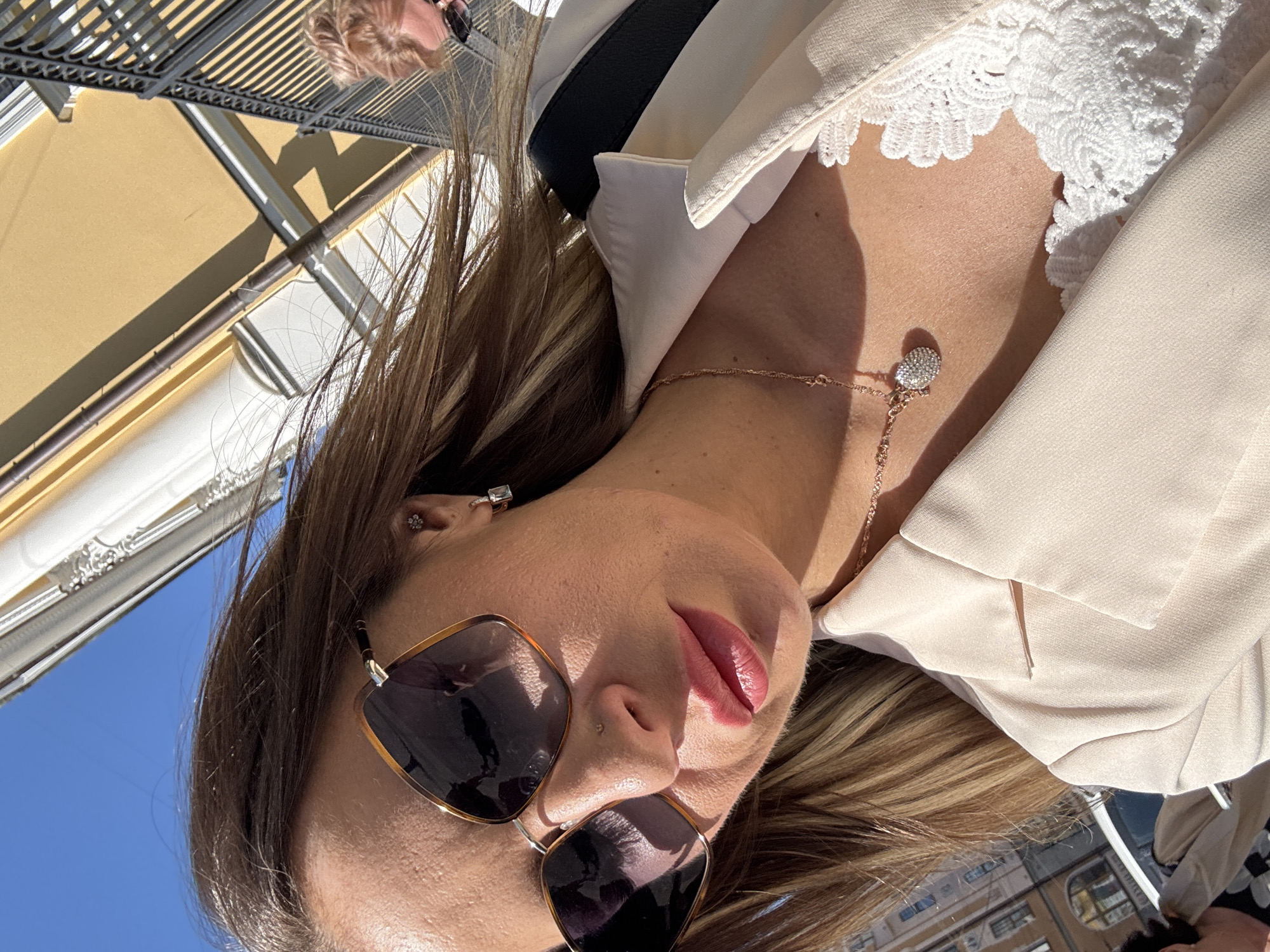 This is a close-up, low-angle, sideways shot of a woman with blonde hair, wearing sunglasses, a beige blazer, and a delicate rose gold necklace with a diamond pendant. She has light pink lipstick on and a small nose ring. The background shows a clear blue sky and part of a building with a yellow facade, possibly indicating an outdoor setting on a sunny day. A portion of a black bag is visible under her blazer, and a lace trim from her inner garment is visible at her neckline. The scene appears to be set in Saint Petersburg, Russia, suggested by the architectural style of the building. The sunlight creates strong shadows, highlighting the woman's face and jewelry. The overall mood is casual and summery. No visible text is present in the image.