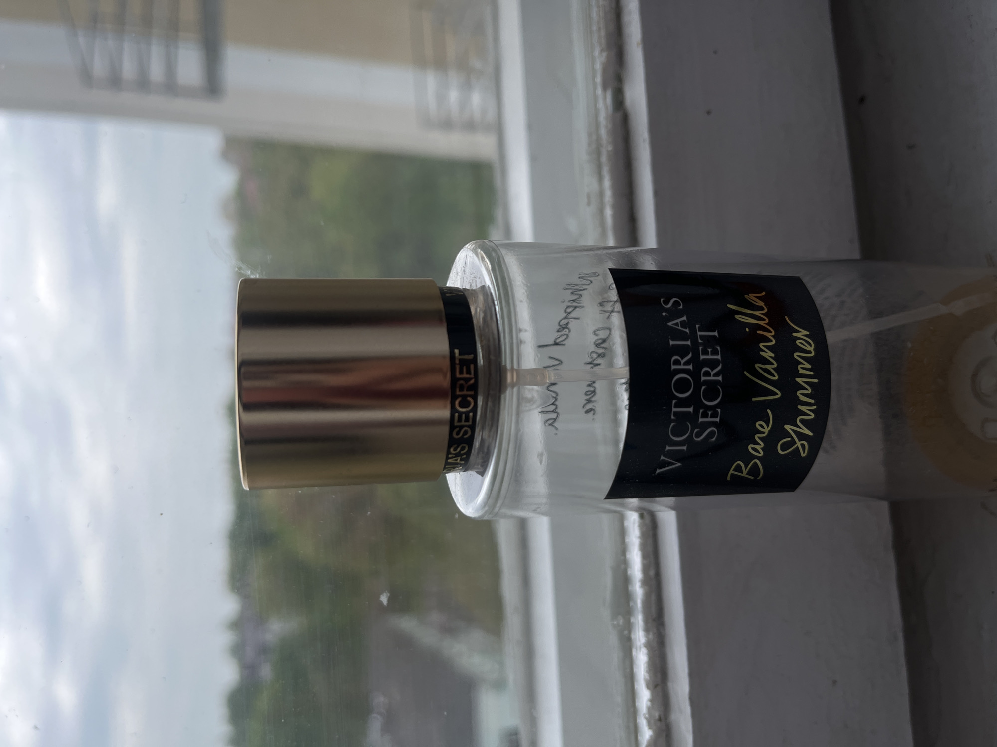 A bottle of Victoria's Secret Bare Vanilla Shimmer fragrance mist is resting on a white windowsill. Outside the window, the sky is overcast and glimpses of greenery are visible. Handwritten text is visible on the bottle above the product label. The location in the city of Maladzyechna, Belarus is not possible to determine from the image.