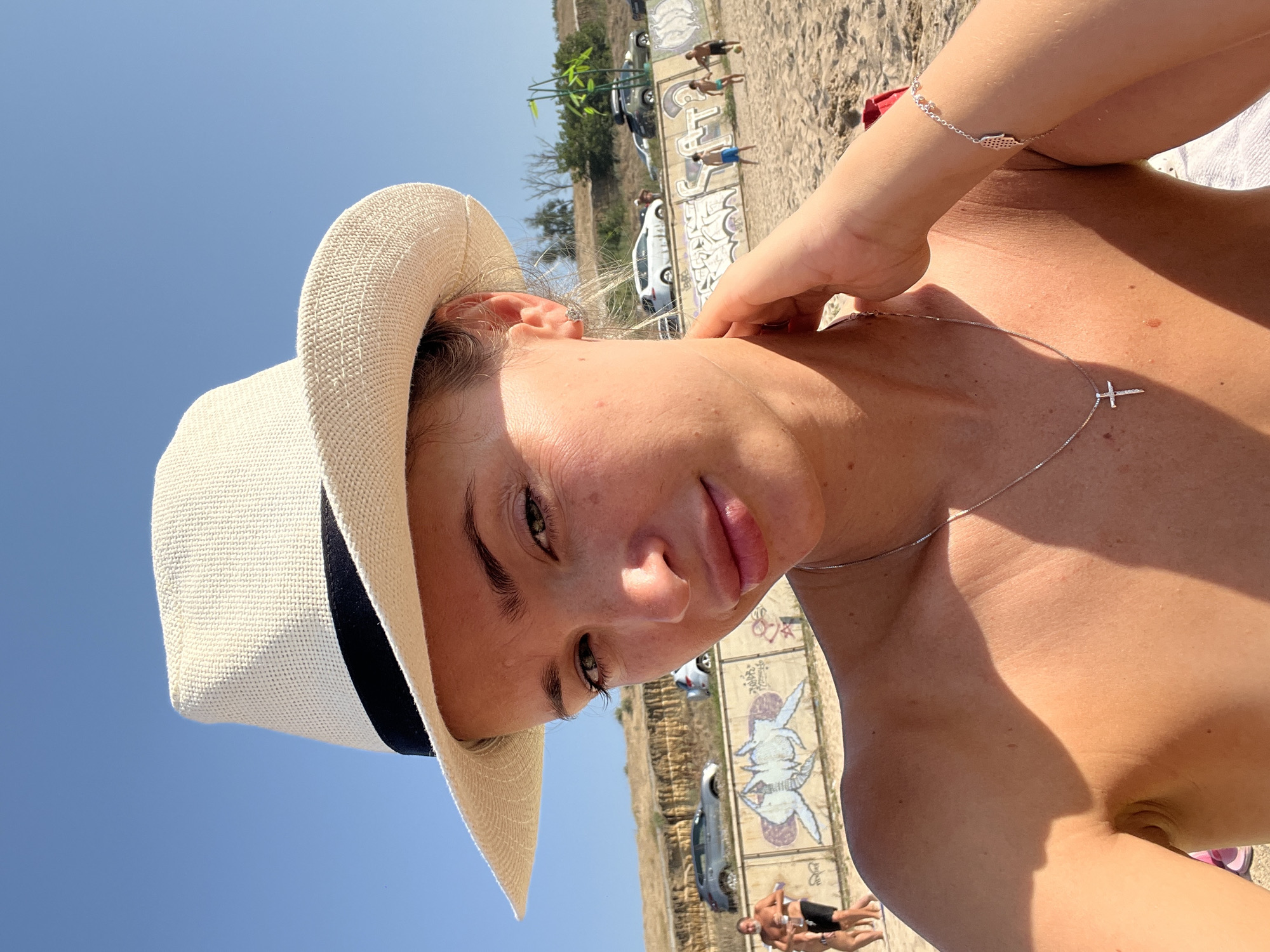 A woman in a white brimmed hat poses for a selfie on a sunny beach. She is wearing a silver necklace with a cross pendant and a silver bracelet on her right wrist. The background shows a sandy beach with people in the distance, some playing soccer. There is a graffiti-covered wall and parked cars on a hill overlooking the beach. The sky is clear blue.