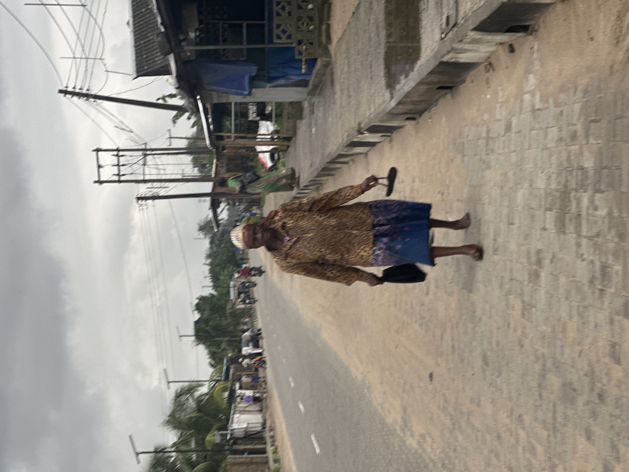 An elderly woman walks along a paved sidewalk next to a road in Badagry, Nigeria. She is wearing a leopard-print shirt and a blue skirt. She is carrying a black bag and a sandal in her hands. The road is mostly empty, with a few motorcycles and cars in the distance. The sky is overcast, and the general atmosphere of the scene is subdued. There are various structures lining the road, including what appear to be shops or market stalls. Utility poles and power lines are visible above the structures. The paved area appears to be a sidewalk next to the road, with a gutter and some sand and debris. The overall scene depicts a typical street view in a Nigerian town under cloudy weather.