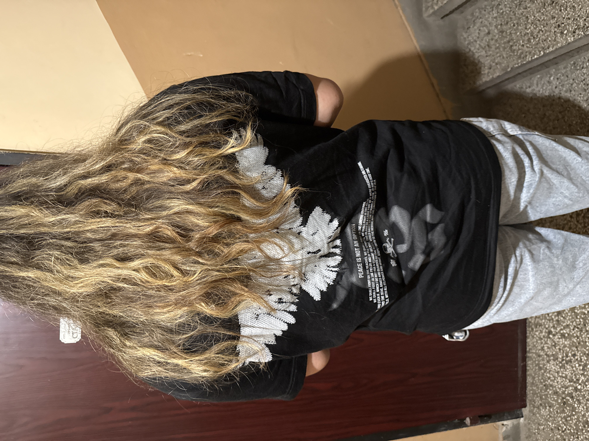 A person with long, wavy, blonde and brown hair stands with their back to the camera, wearing a black t-shirt with a white graphic of a bird's wings and text. The text on the shirt reads "PEACE IS NOT AN OPTION" and continues with smaller text that is partially legible. The person is also wearing light grey sweatpants. They are standing indoors, in a hallway with a dark red door on the left and a light tan wall on the right. The floor is a speckled concrete material, and stairs are visible on the right side of the frame. There is a small white rectangular sticker on the dark red door. The lighting is artificial. The scene is consistent with a residential building hallway in Plovdiv, Bulgaria.