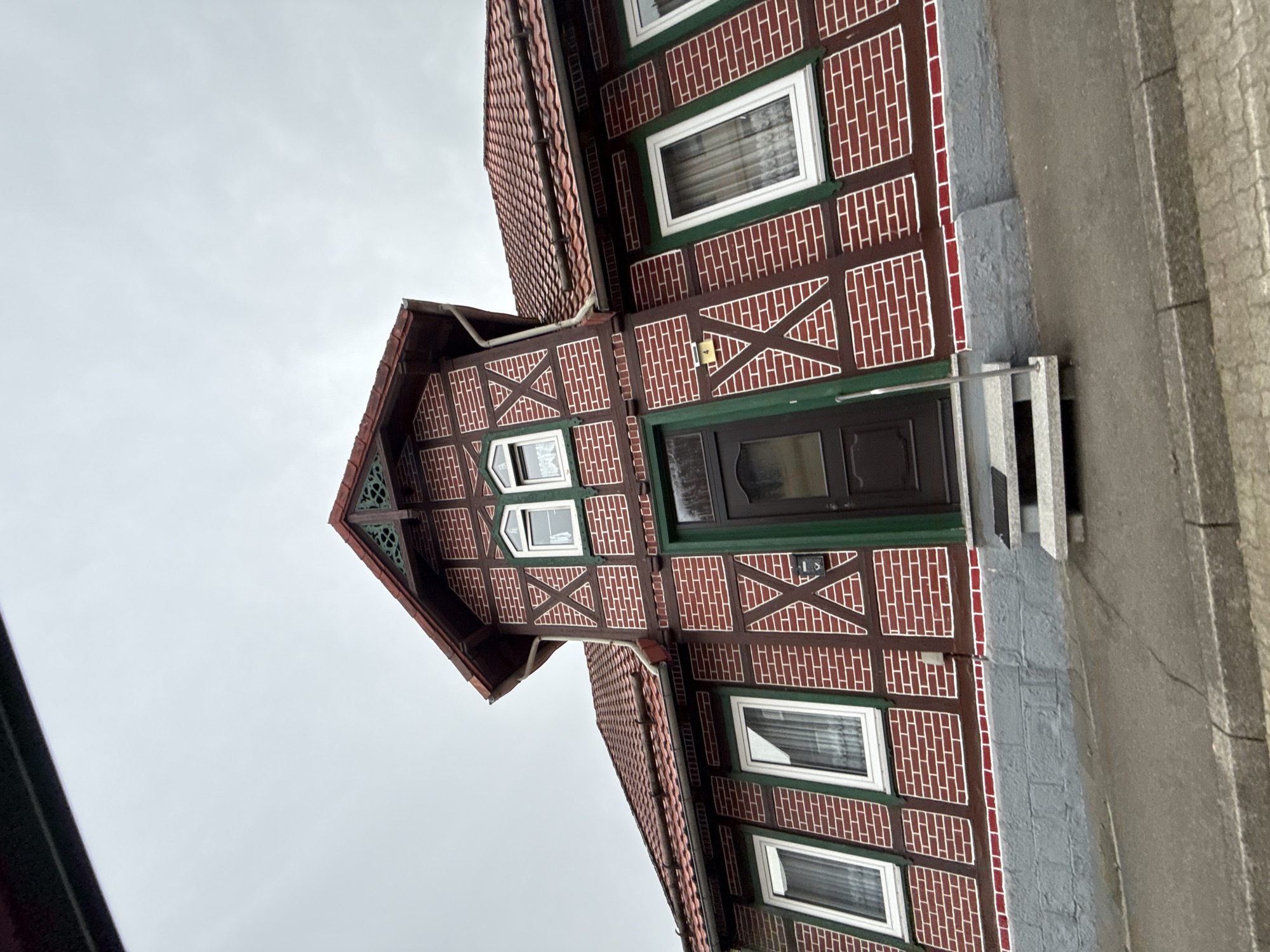 A front-facing, eye-level, medium shot captures a traditional half-timbered house in the city of Bad Sachsa, Germany. The building features a red tile roof, red brick walls with dark brown timber framing outlined in white, and green window frames. Multiple windows with white frames and curtains are visible on the facade. A dark brown door with a green frame is centrally located, accessed by a small set of grey, stone-like steps. The lower part of the house is a grey stone foundation. The scene is set against an overcast, grey sky, with a paved walkway and curb in the foreground. The overall impression is of a residential building in a European town.