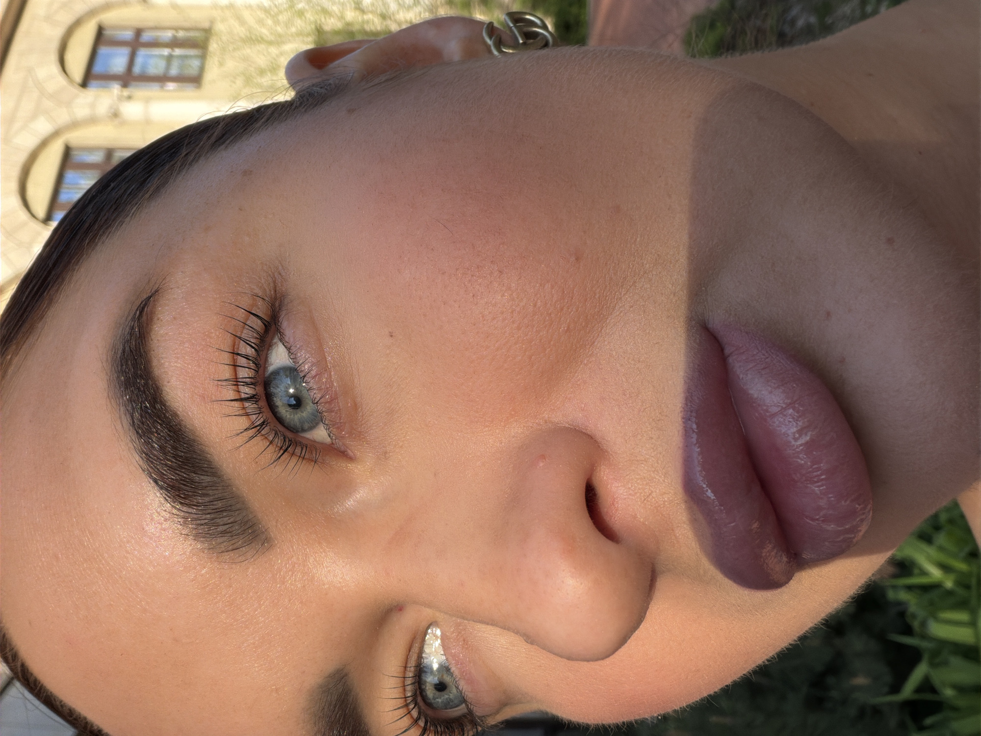 The image shows a close-up of a woman's face, angled upward. Her features include blue eyes with long eyelashes, defined eyebrows, a nose, and lips colored with a deep purple lipstick. A silver ring is visible in her ear. In the background, out of focus, is the architecture of a building, hinting at an urban location with arched windows. Based on the architectural style, particularly the arched windows and overall design, this could be a location in Moscow, Russia. There is also some greenery that suggests the scene might be in a park or garden. The lighting indicates that the photo was taken outdoors, possibly in direct sunlight.