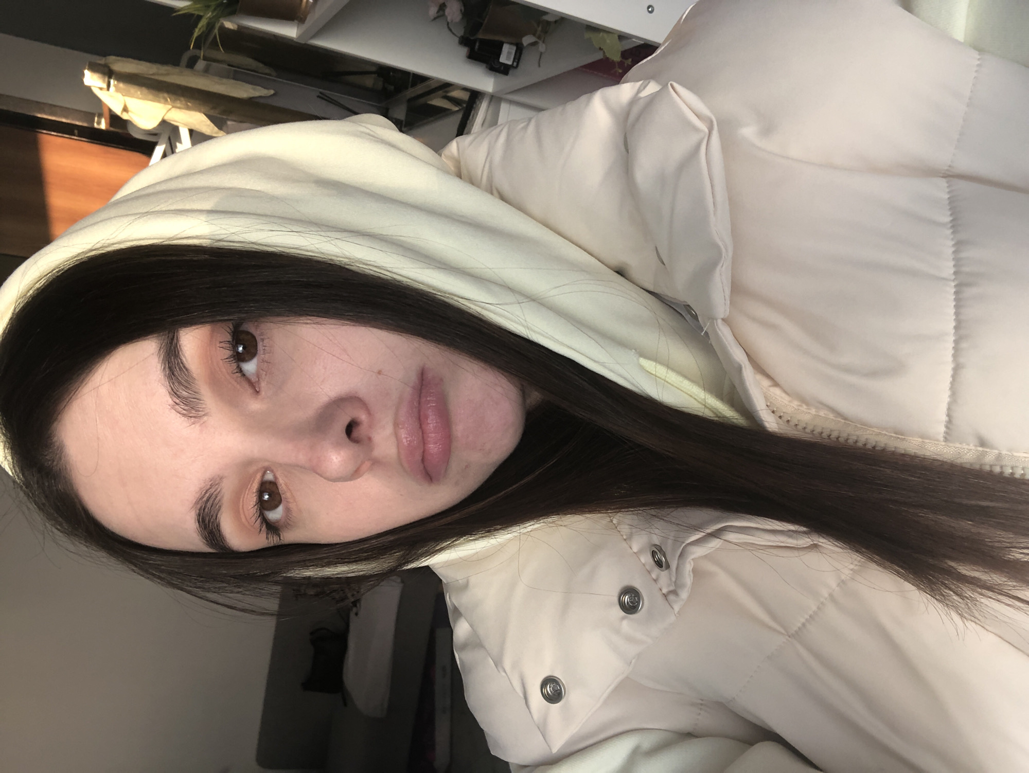 A close-up, selfie-style shot captures a young woman with long, dark brown hair. She is wearing a light-colored, possibly cream or beige, puffy jacket with a hood. The hood is up, framing her face. Her skin appears fair, and her features include prominent eyebrows, brown eyes, a nose with a slight bump, and full lips. The background suggests an interior space, with shelves and various objects visible in the blurred background, including what appears to be a light fixture and some greenery. There is no identifiable location in the city of Chaikovsky, Russia.