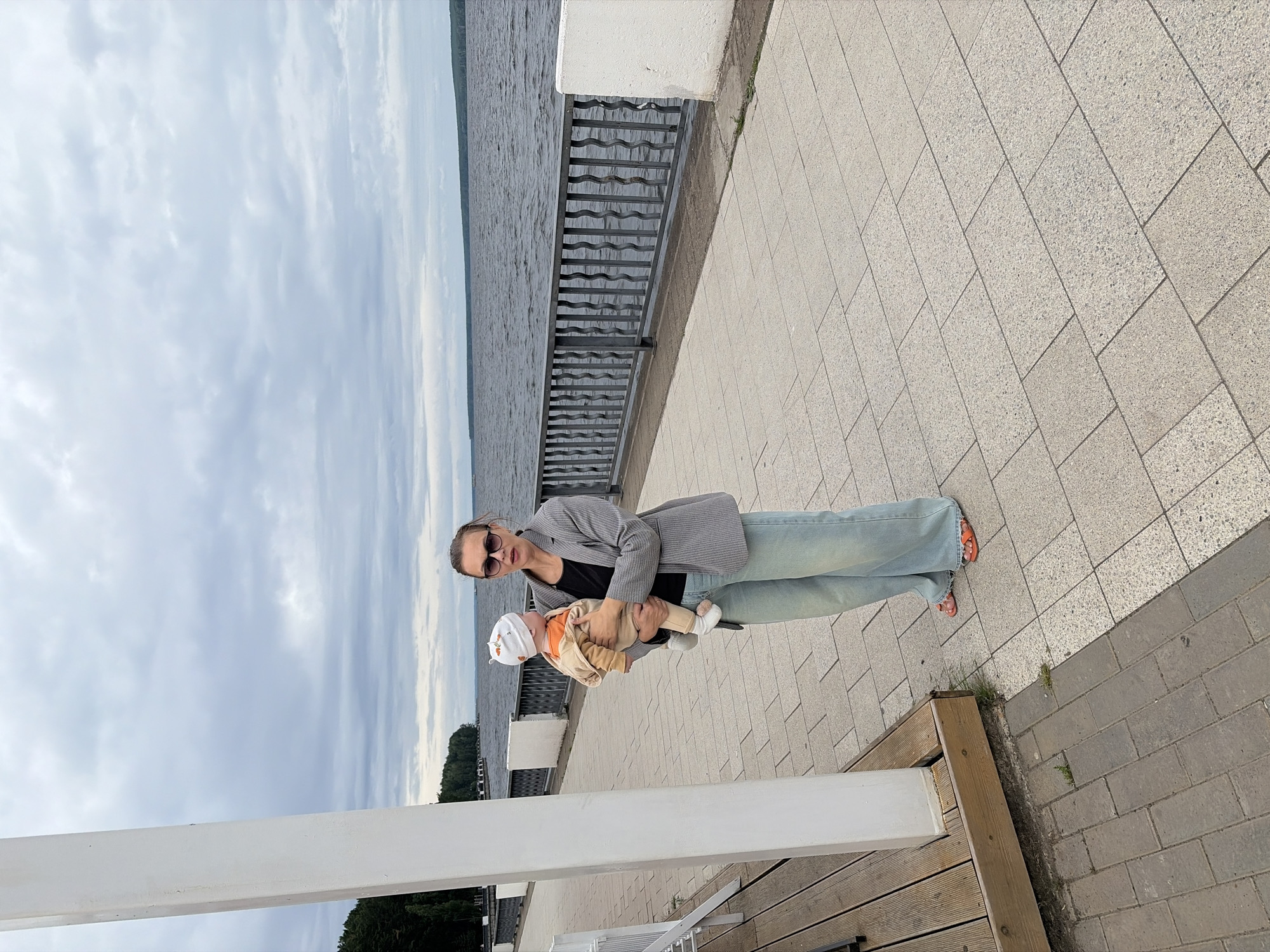 A woman is holding a baby while standing on a paved walkway next to a body of water. The woman is wearing a grey pinstripe blazer over a black shirt, light blue jeans, and orange sandals. She is also wearing sunglasses. The baby is wearing a white hat with an orange pattern and an orange and tan outfit. The background shows a body of water with a cloudy sky. The walkway is made of light-colored square tiles, and there is a black metal railing separating the walkway from the water. A wooden bench is visible to the right of the walkway. The scene appears to be outdoors during the daytime, with a cloudy sky. The location is Chaikovsky, Russia.